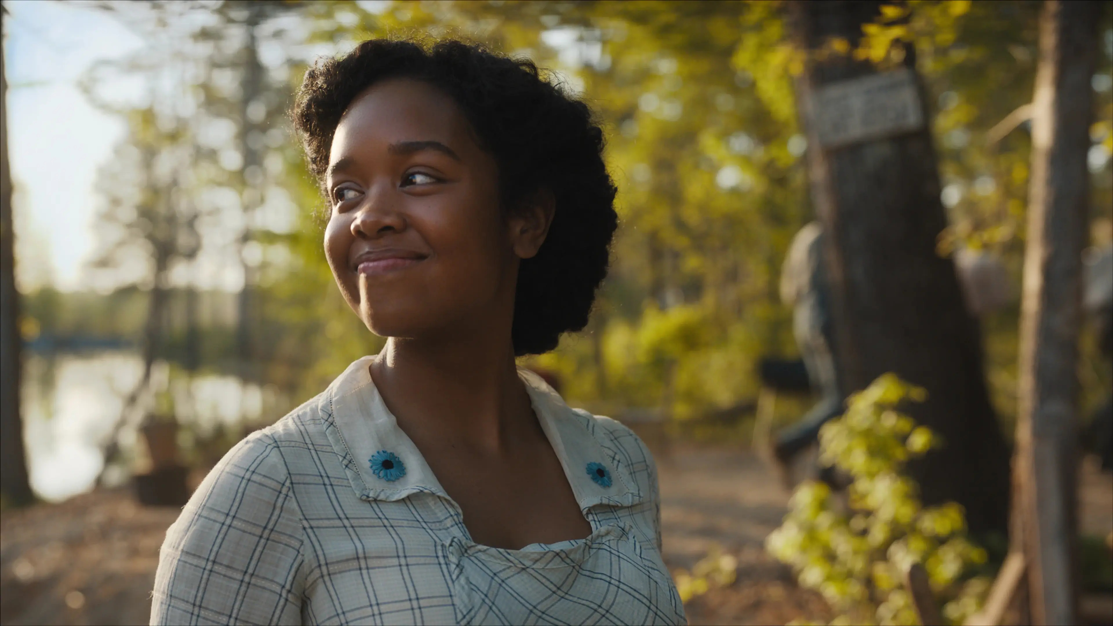 A woman in a plaid dress with blue floral trim smiles outdoors in The Color Purple (2023).