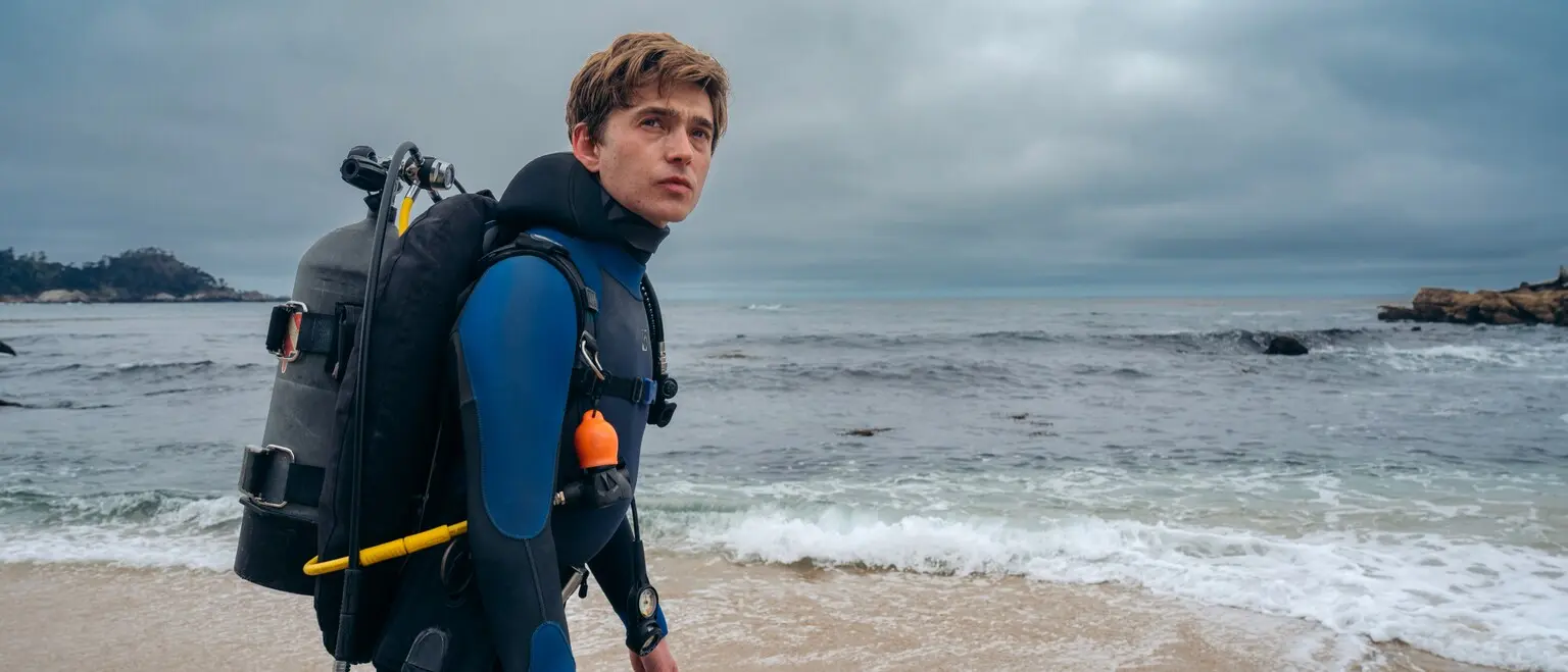 Austin Abrams as Jay Gardiner stands in scuba gear beside the ocean.