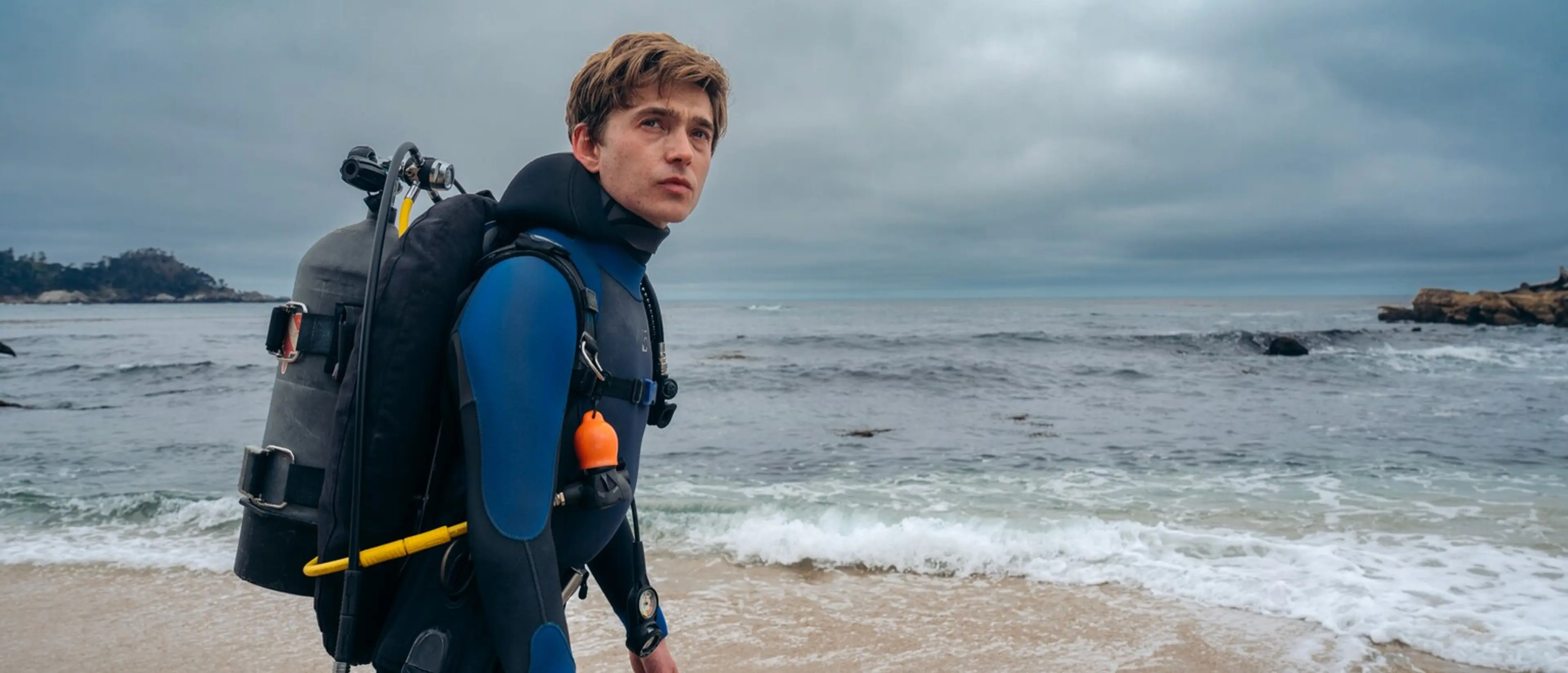 Austin Abrams as Jay Gardiner stands in scuba gear beside the ocean.