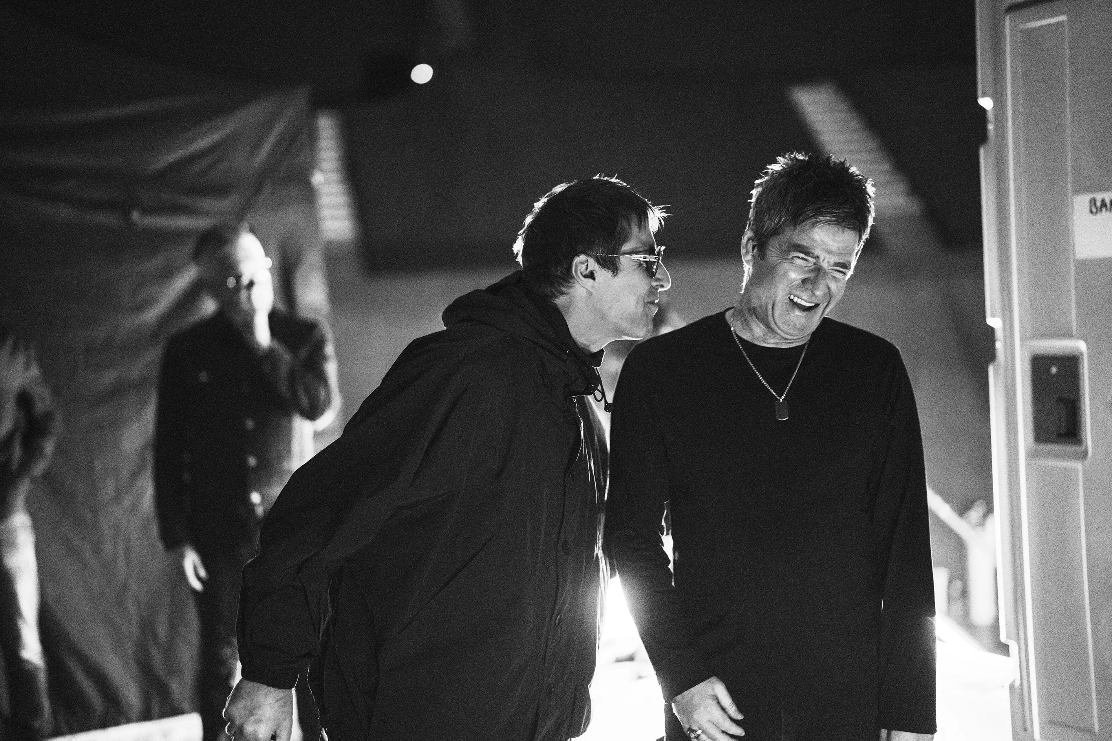 Liam Gallagher leans toward a smiling Noel Gallagher in a black-and-white backstage still.