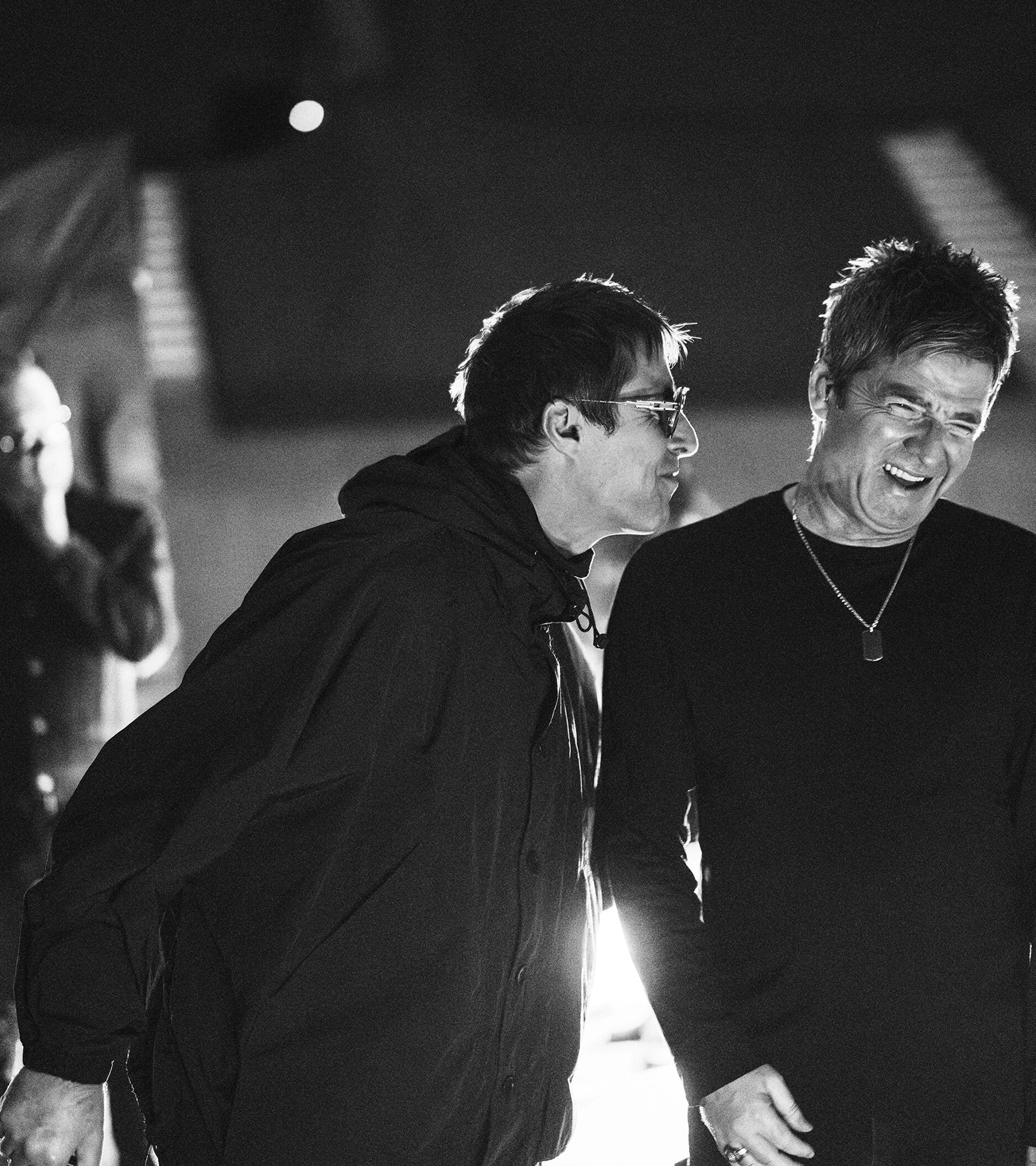 Liam Gallagher leans toward a smiling Noel Gallagher in a black-and-white backstage still.