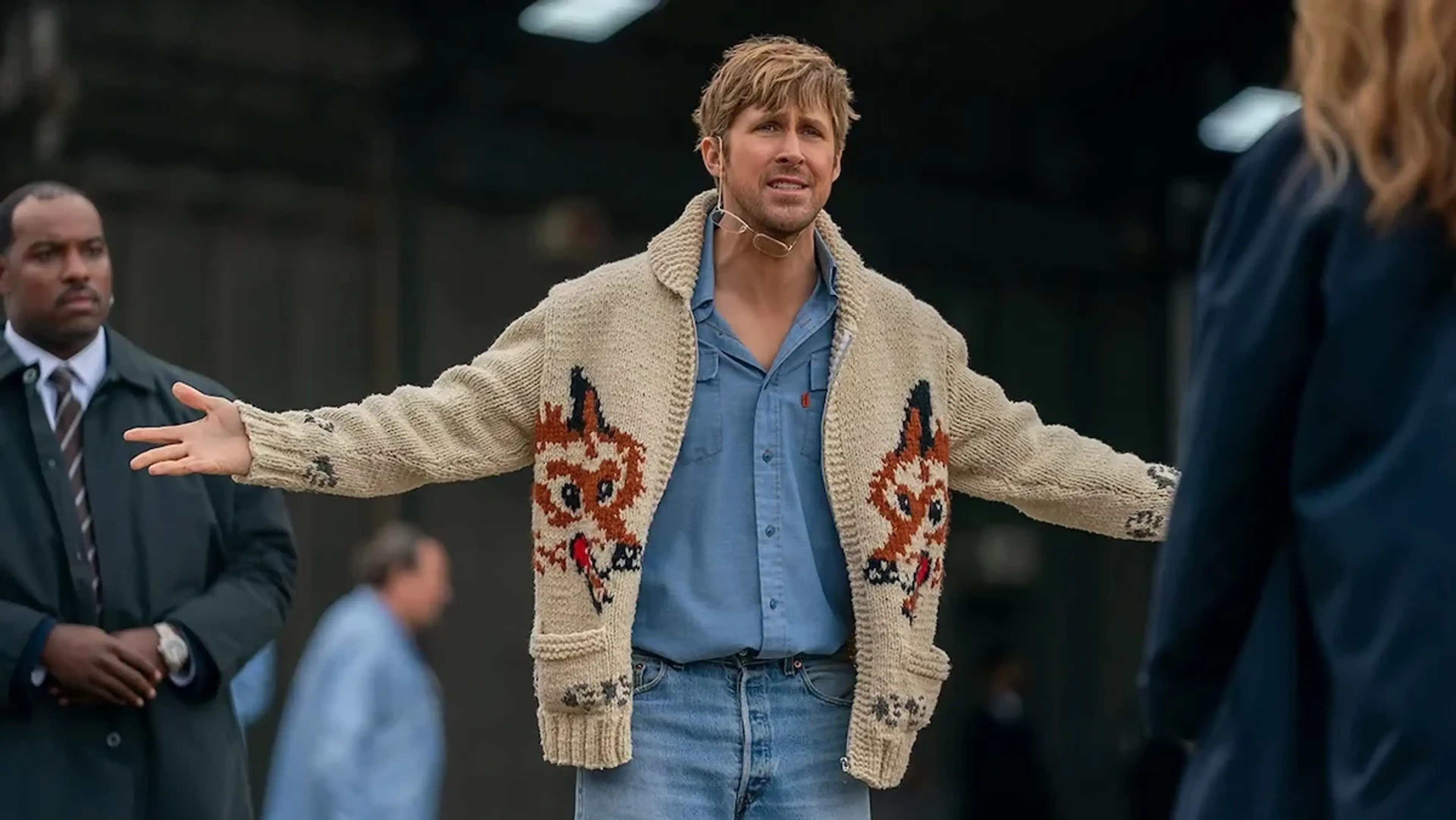 Ryland Grace spreads his arms while wearing a cream cardigan and a blue shirt.