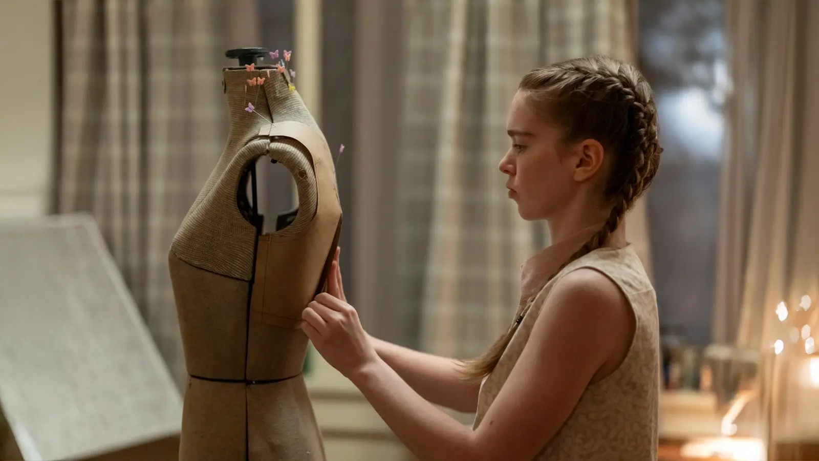 Summer H. Howell as Carrie White puts a pin into a dress form in a softly lit room.