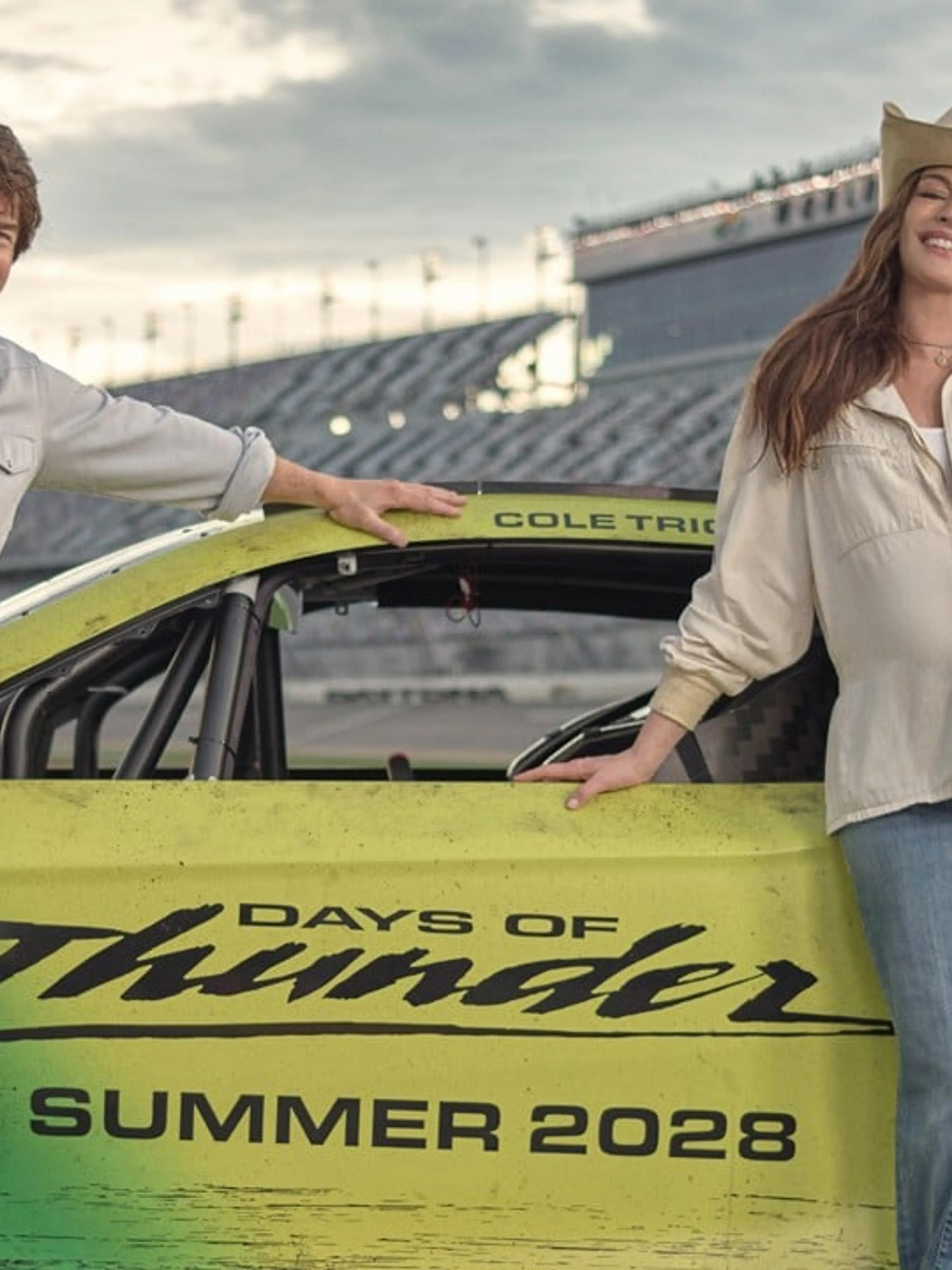 Tom Cruise and Anne Hathaway stand beside the green-and-yellow Days of Thunder race car at Daytona International Speedway.