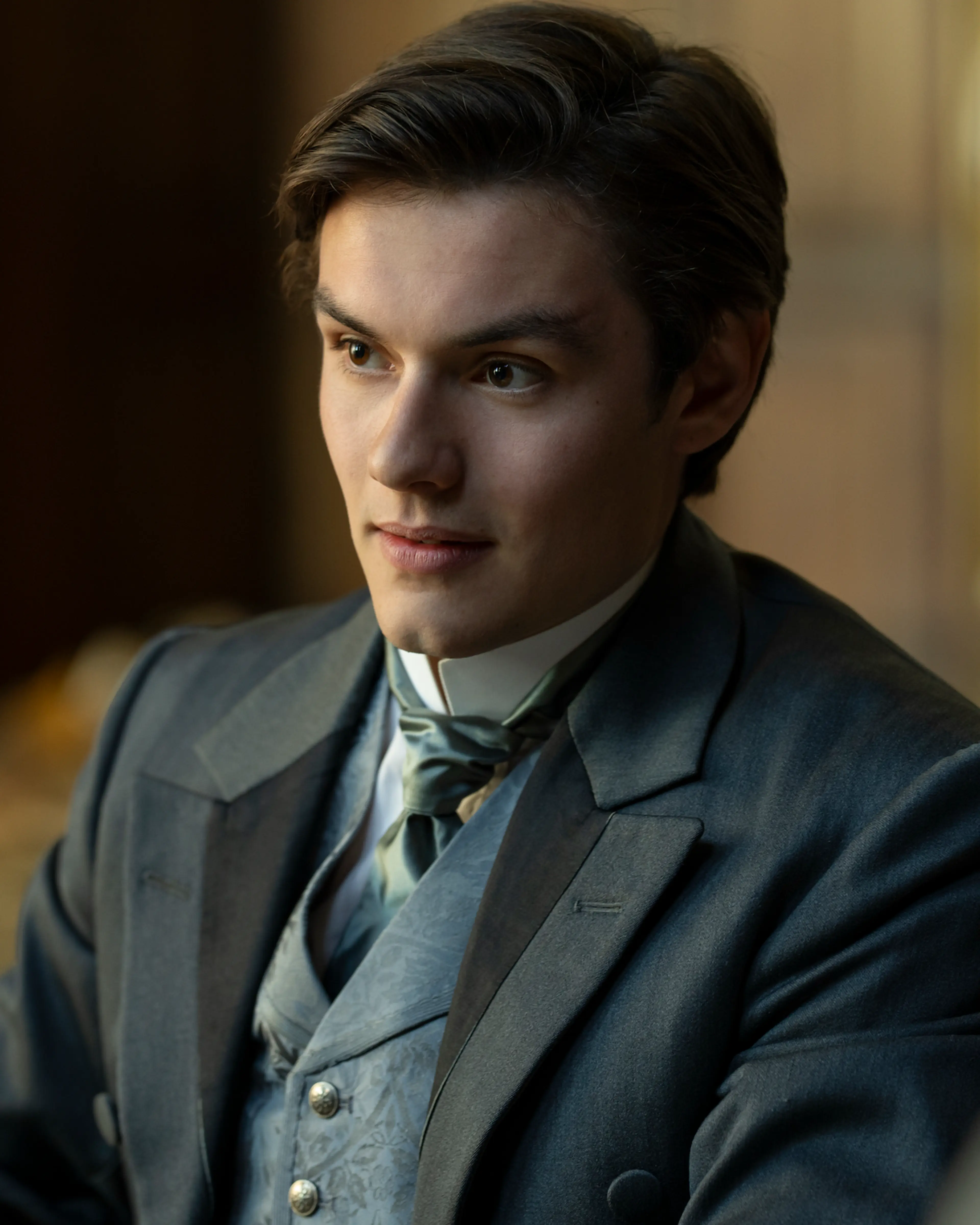 Louis Partridge as Tewkesbury, wearing a blue suit and green necktie.