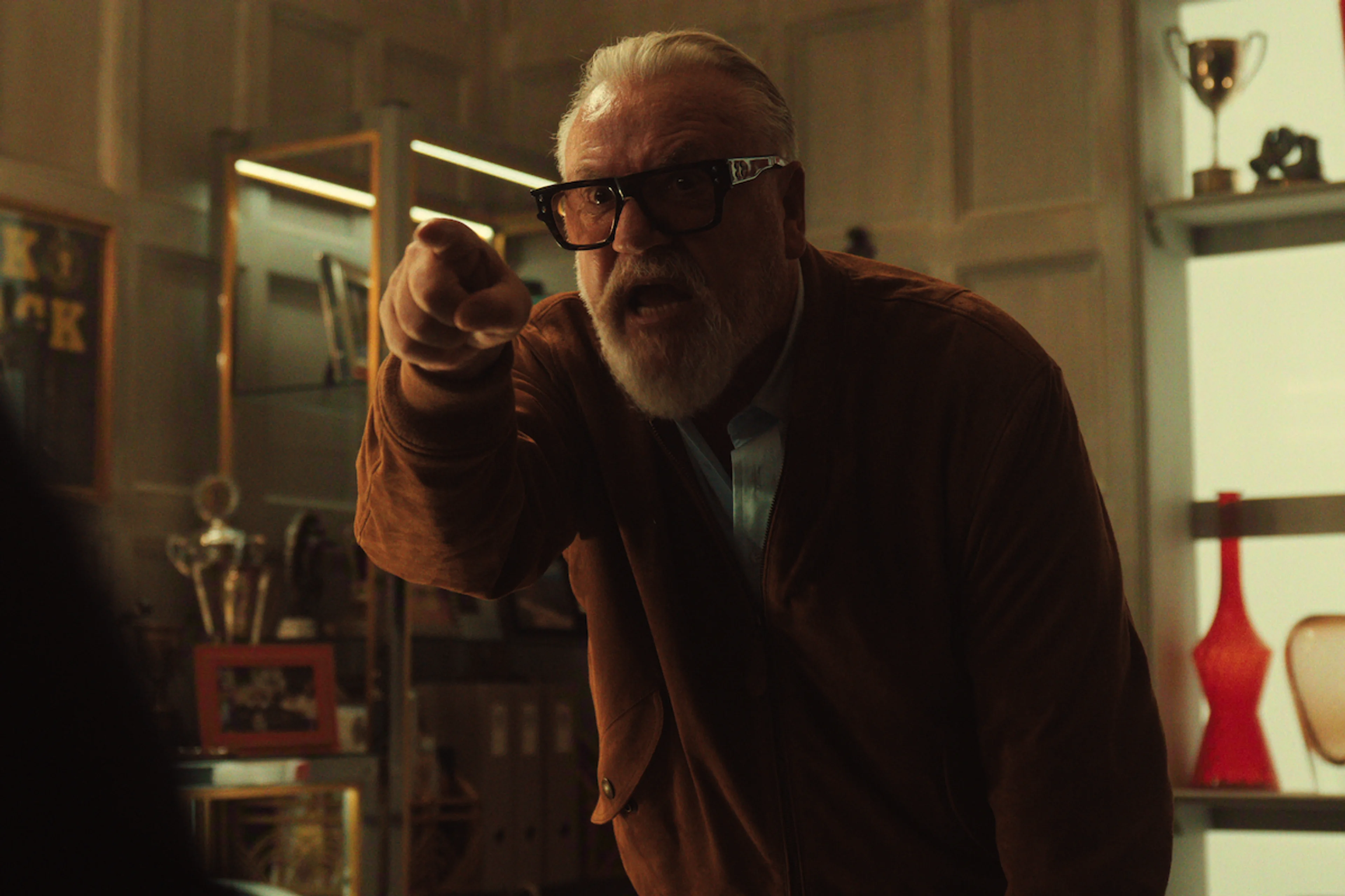 Ray Winstone as Bobby Glass points angrily across a room containing shelves and trophies.