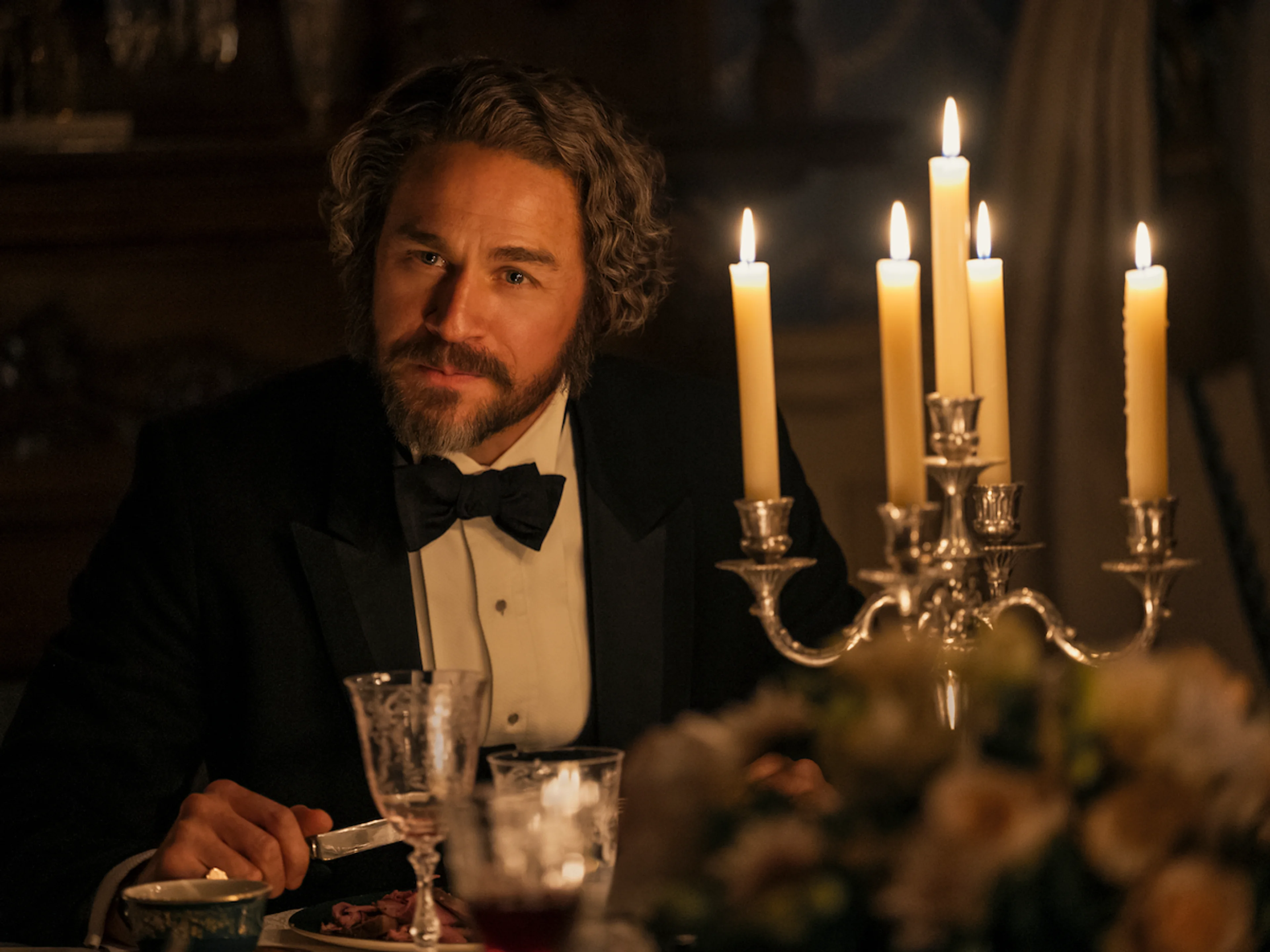 Charlie Hunnam as Andrew Borden sits at a table with food, flowers and candles.