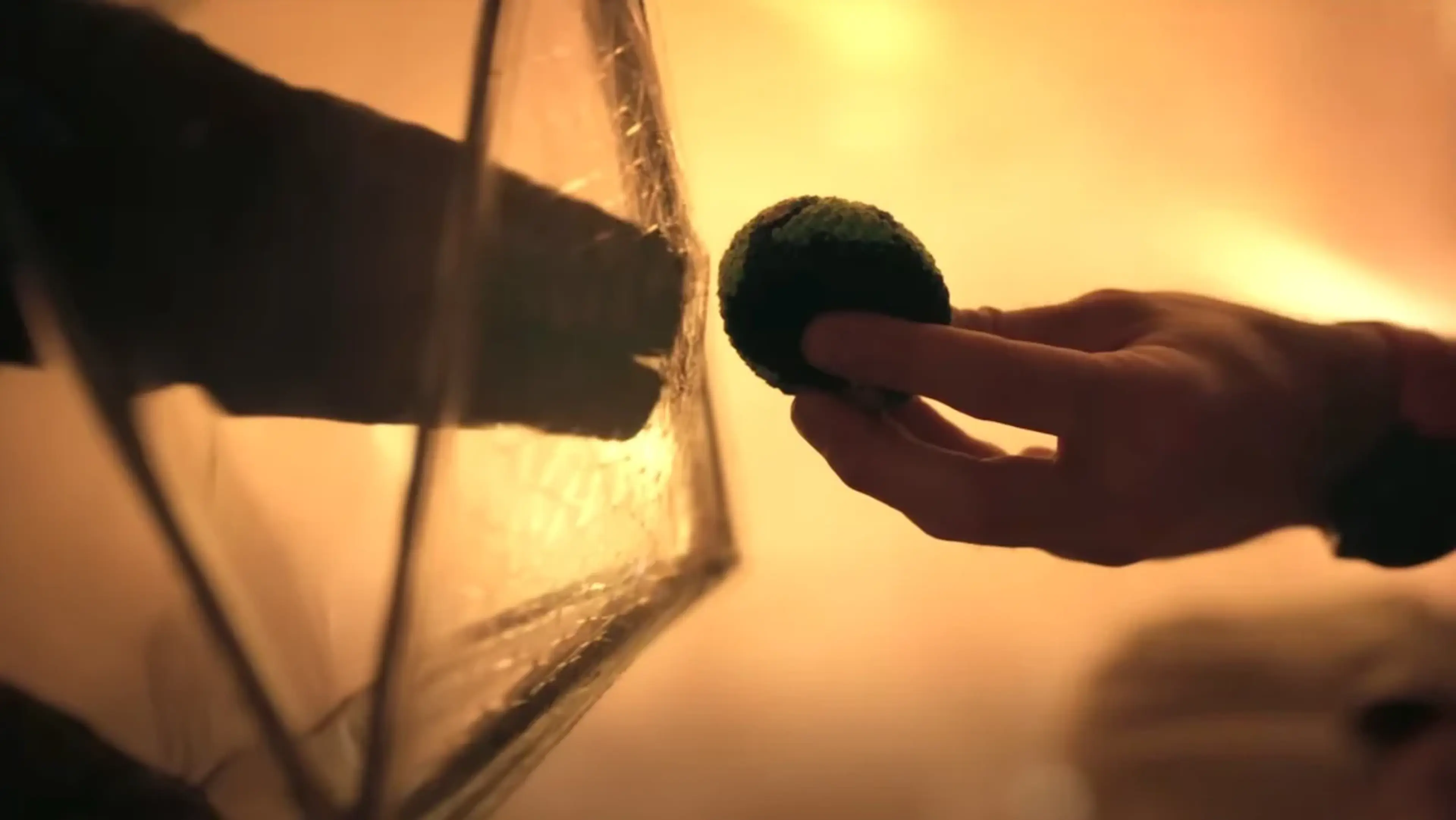 A human hand holds a knitted ball beside a rough alien hand separated by a transparent barrier.