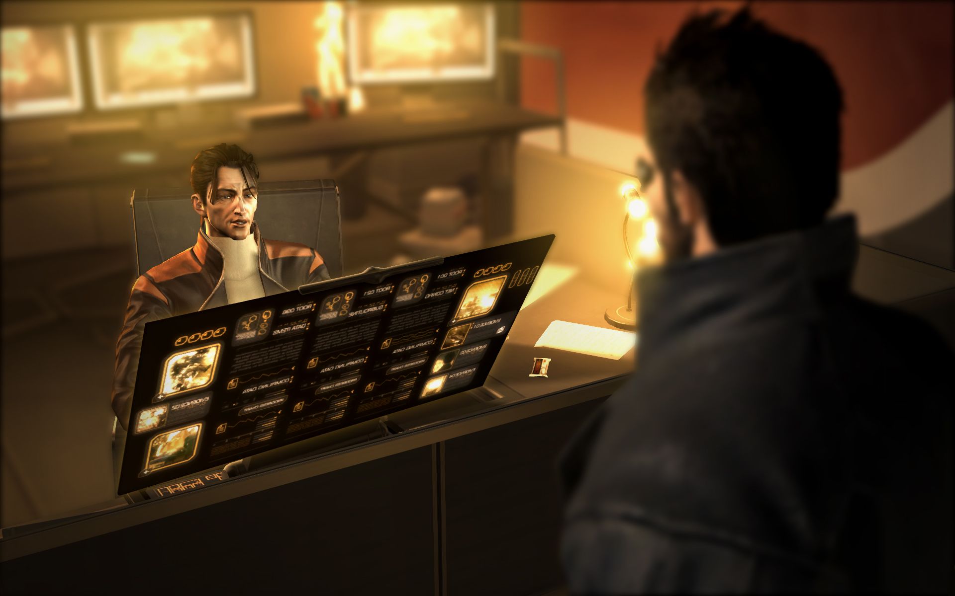 Frank Pritchard sits behind a desk and a bright workstation display, speaking with Adam Jensen.