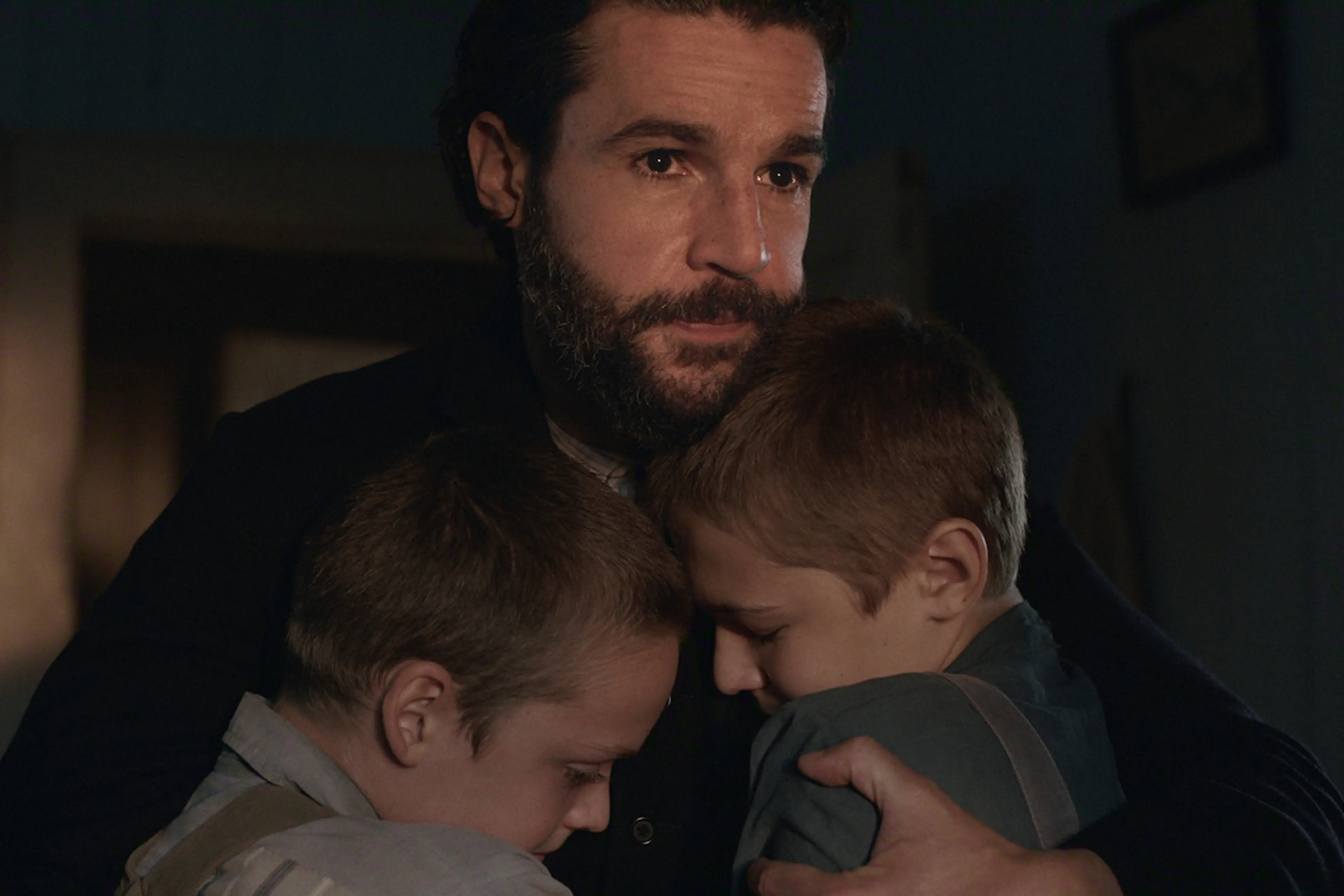 A bearded father holds two young boys close inside a dimly lit home in East of Eden.