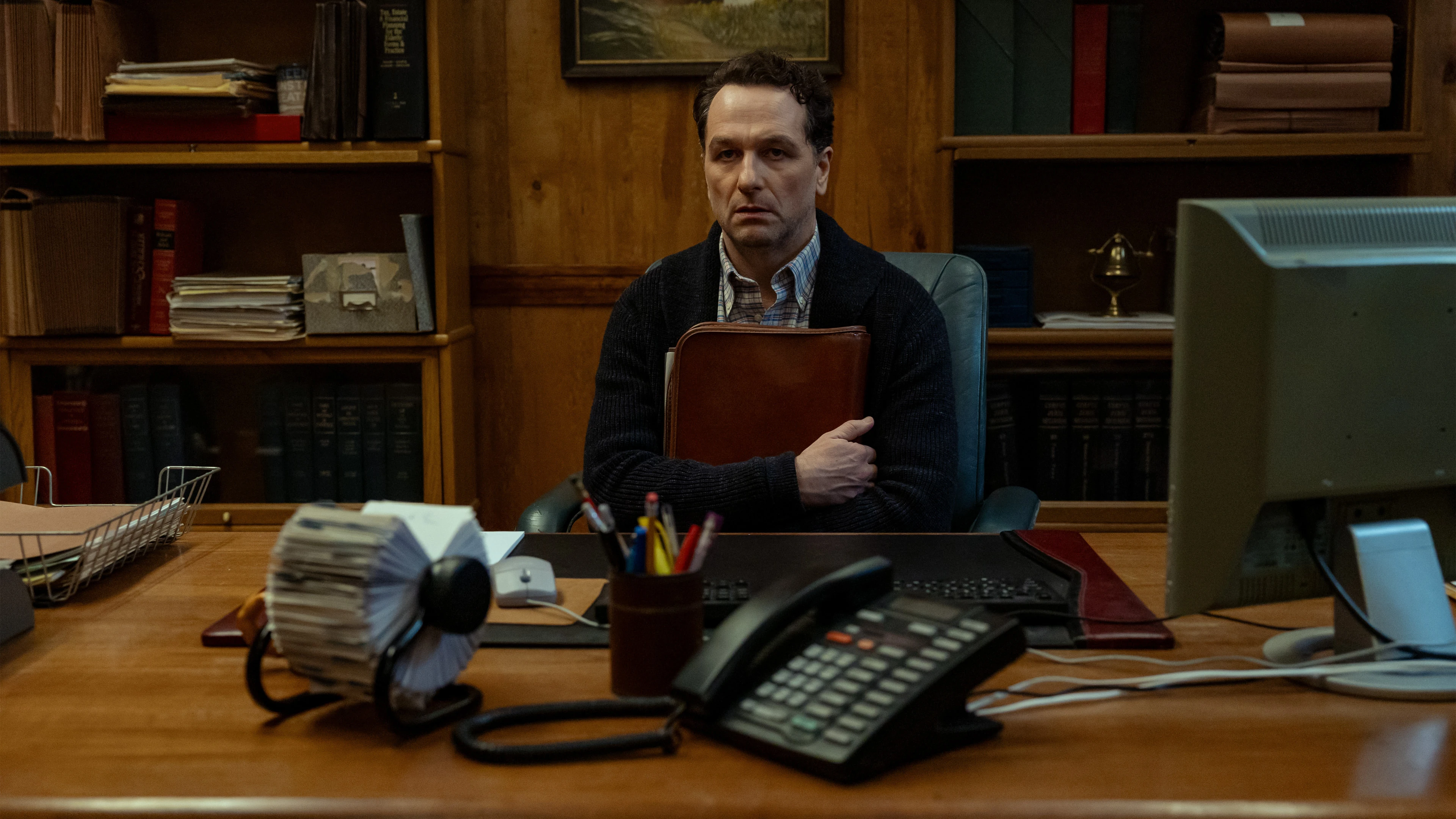 Matthew Rhys as Mayor Tom Loftis sits at his desk, holding a leather folder.