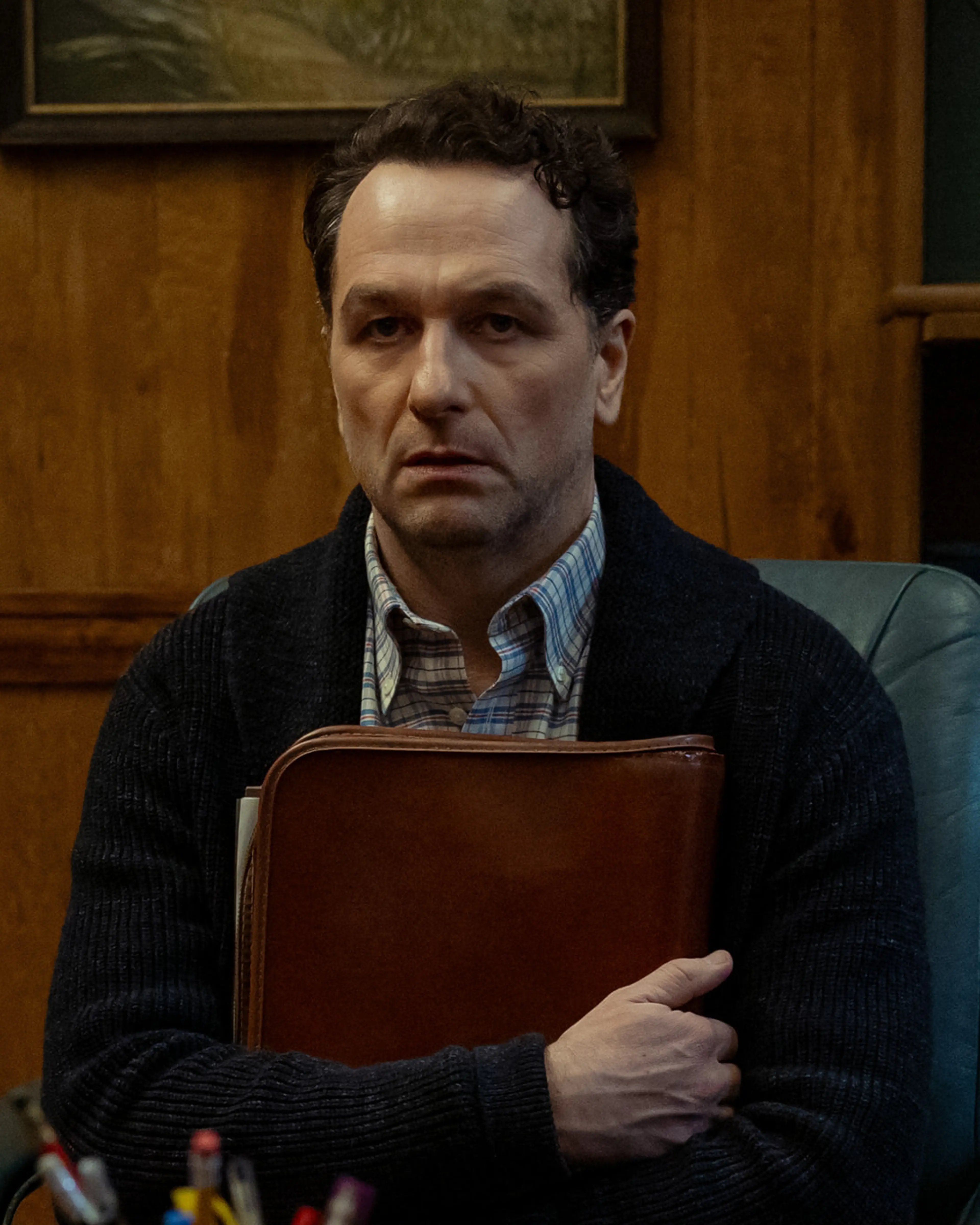 Matthew Rhys as Mayor Tom Loftis sits at his desk, holding a leather folder.