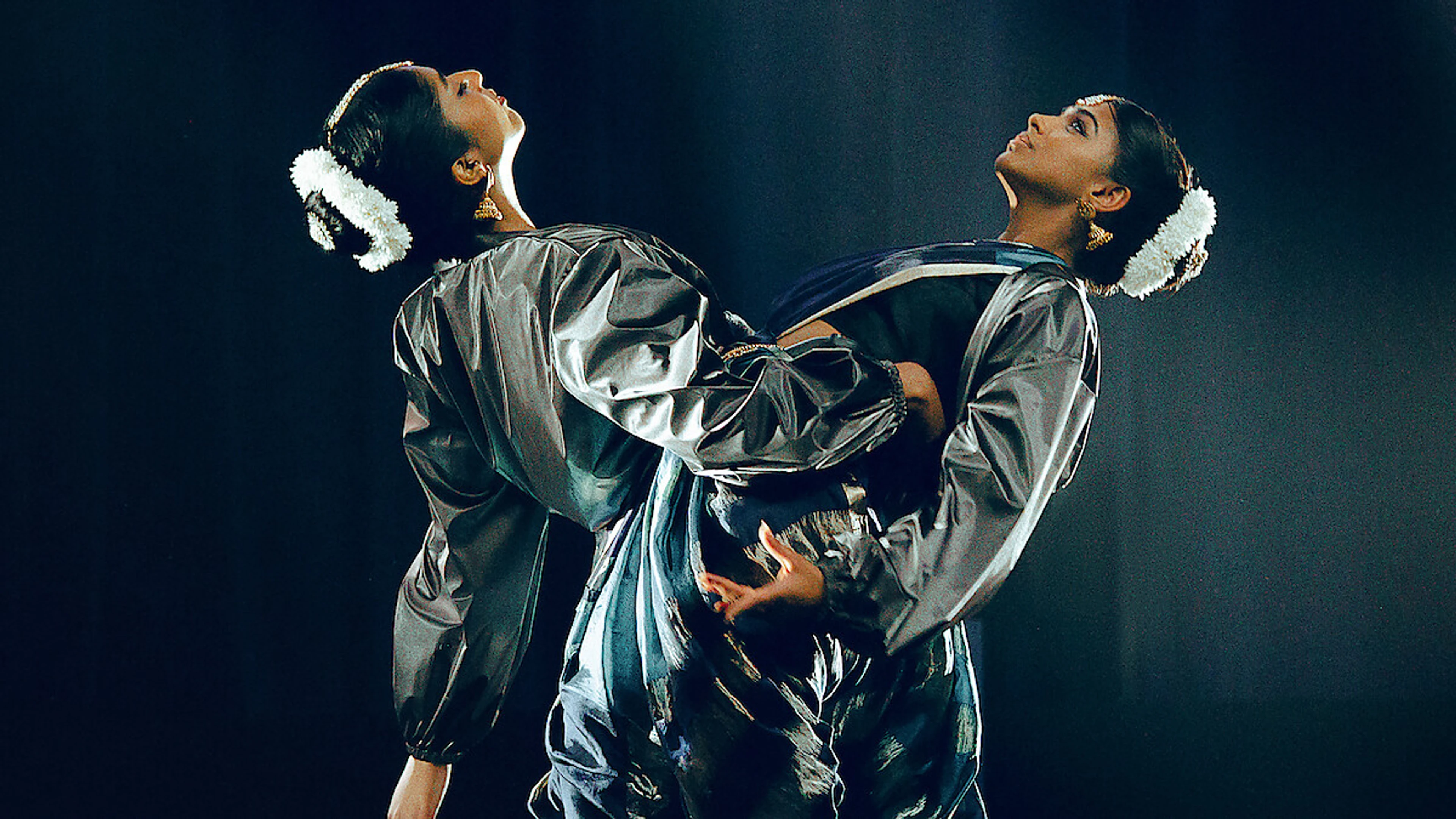 Two dancers in silver costumes lean back to back under stage lights.