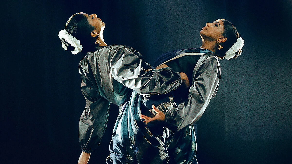 Two dancers in silver costumes lean back to back under stage lights.