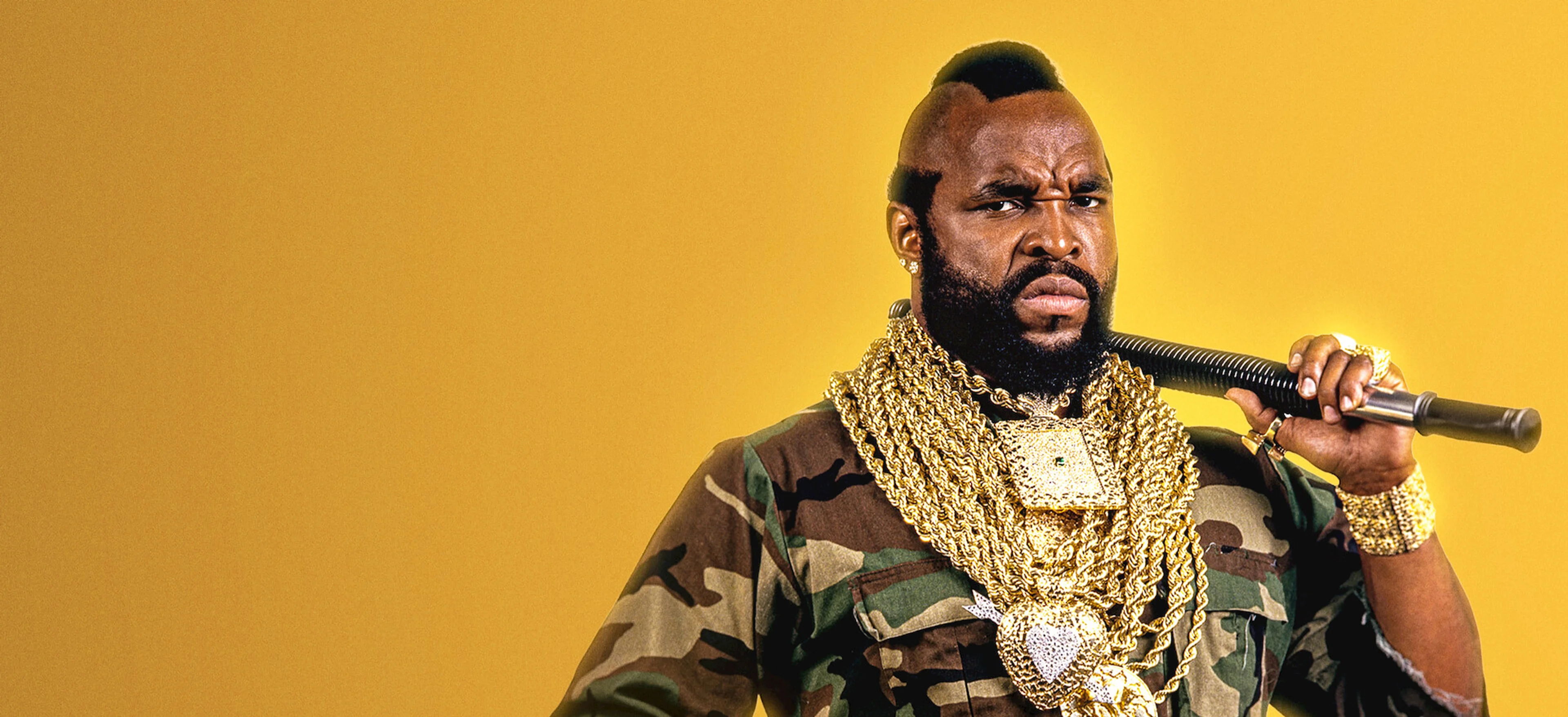 Mr. T wears camouflage and gold chains while holding a baton against a yellow background.