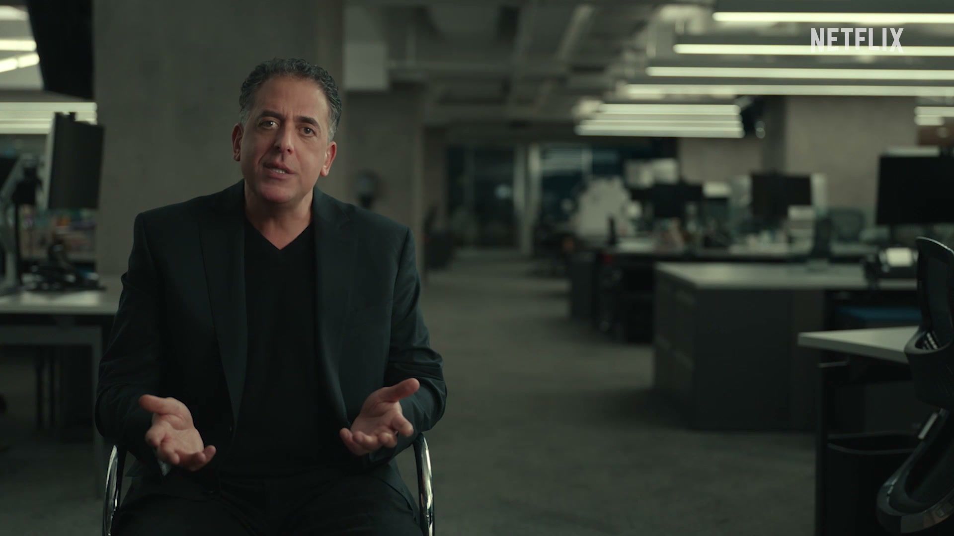 A dark-haired man in a black jacket sits in an office and speaks to camera.