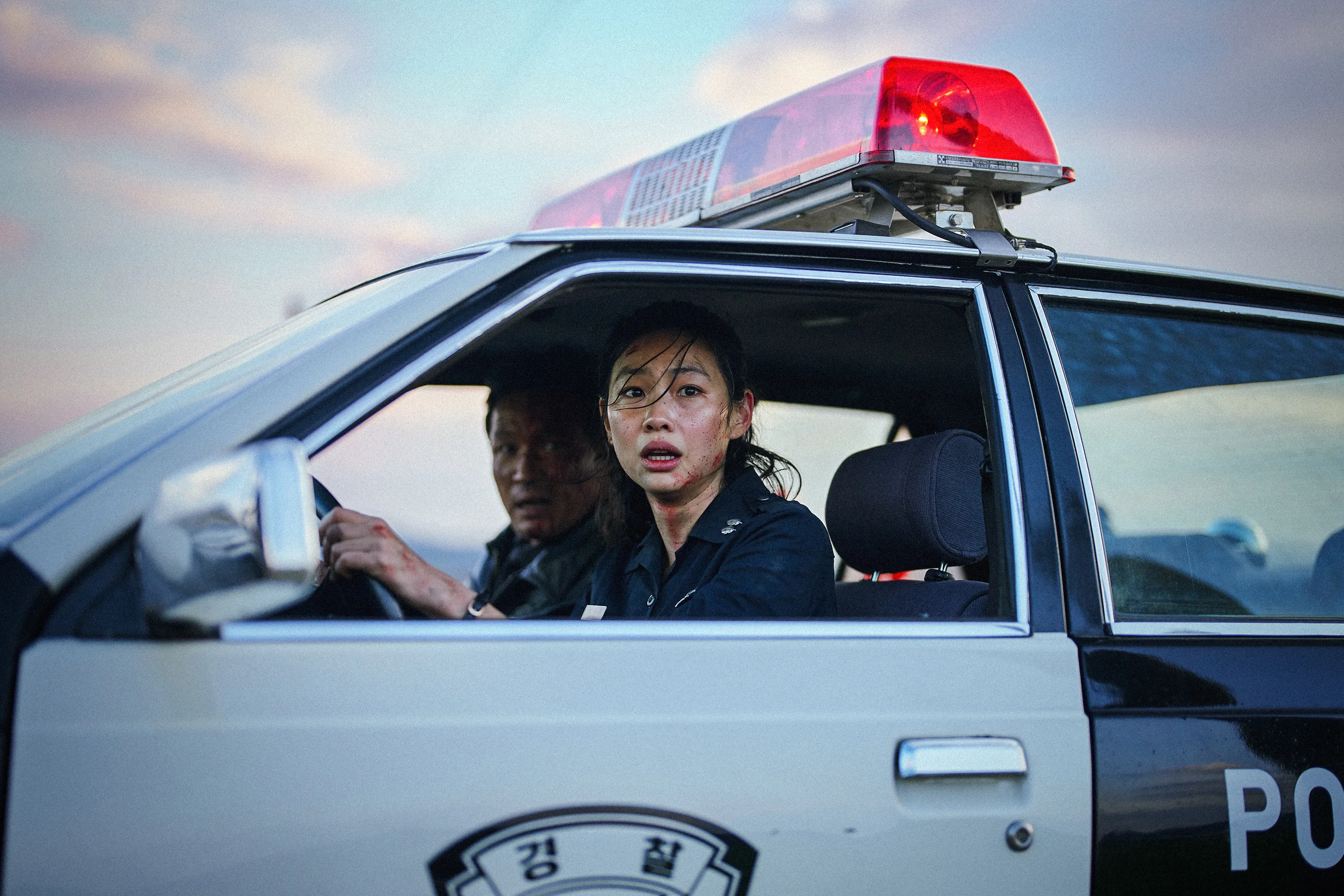 Bum-seok and Sung-ae sit in a police car with a red roof light.