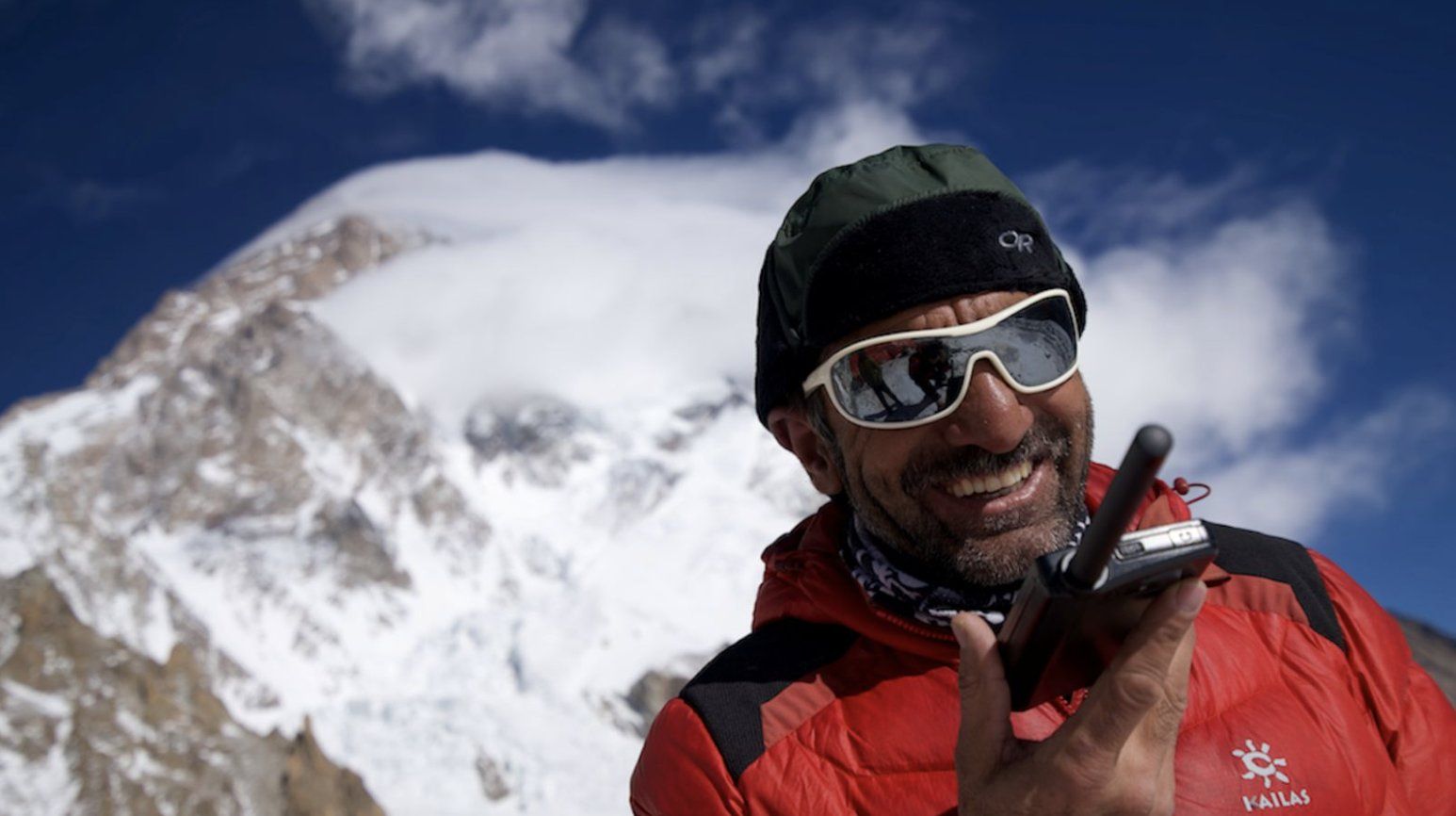 A mountaineer in red winter clothing smiles while holding a radio beneath snowy peaks.