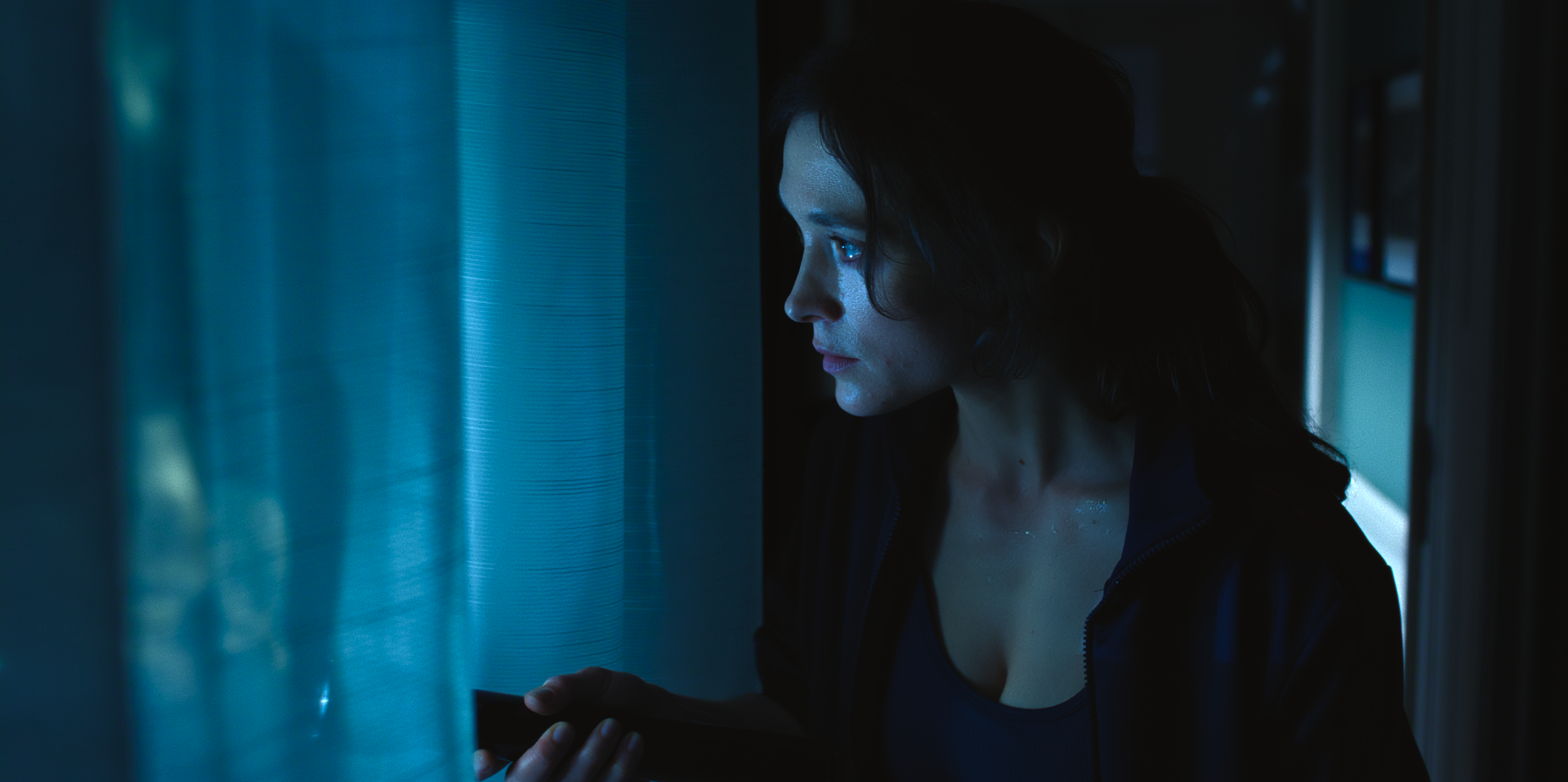 Friederike Becht as Lisa Konrad looks through blue-lit curtains.