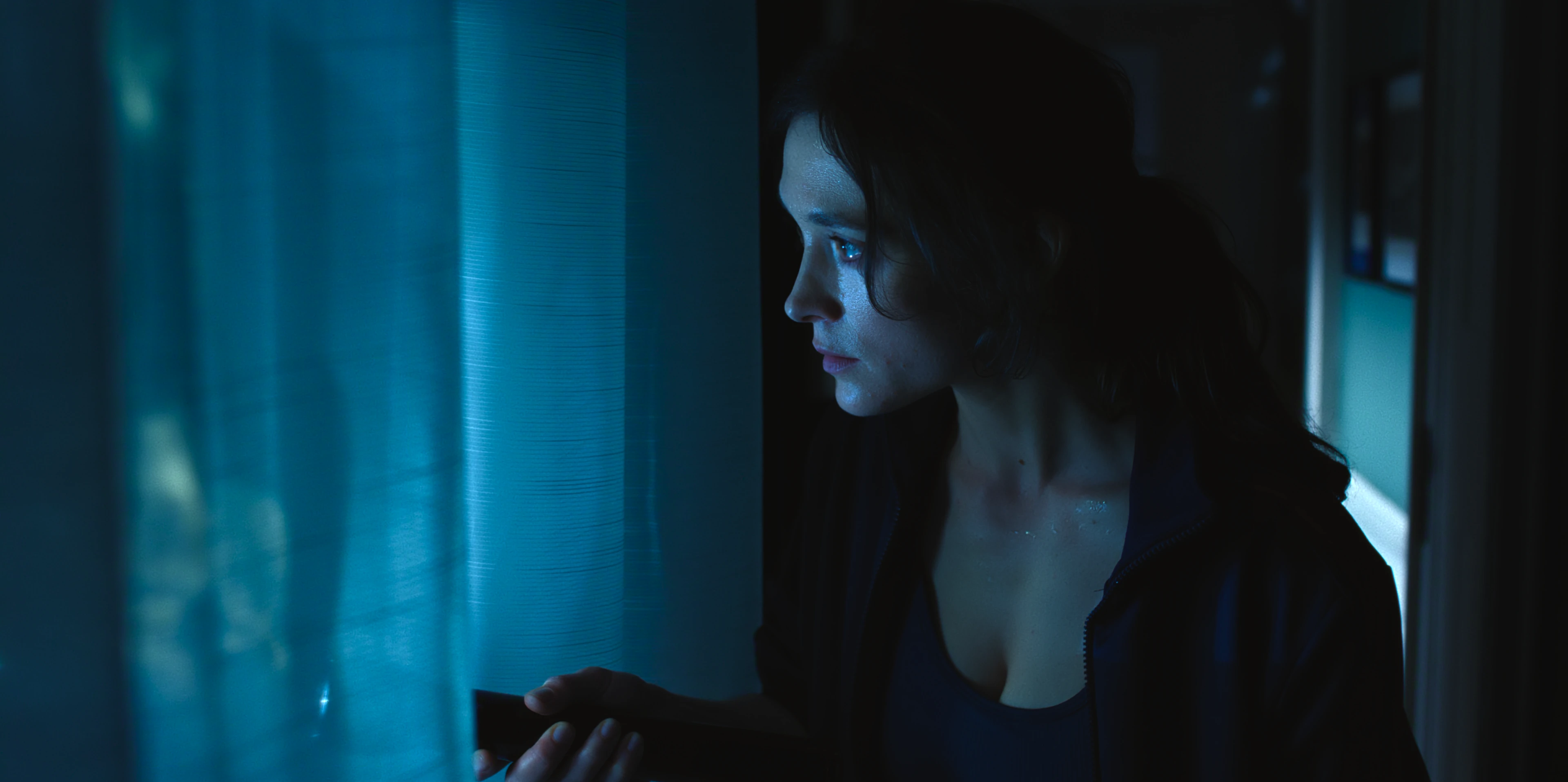 Friederike Becht as Lisa Konrad looks through blue-lit curtains.