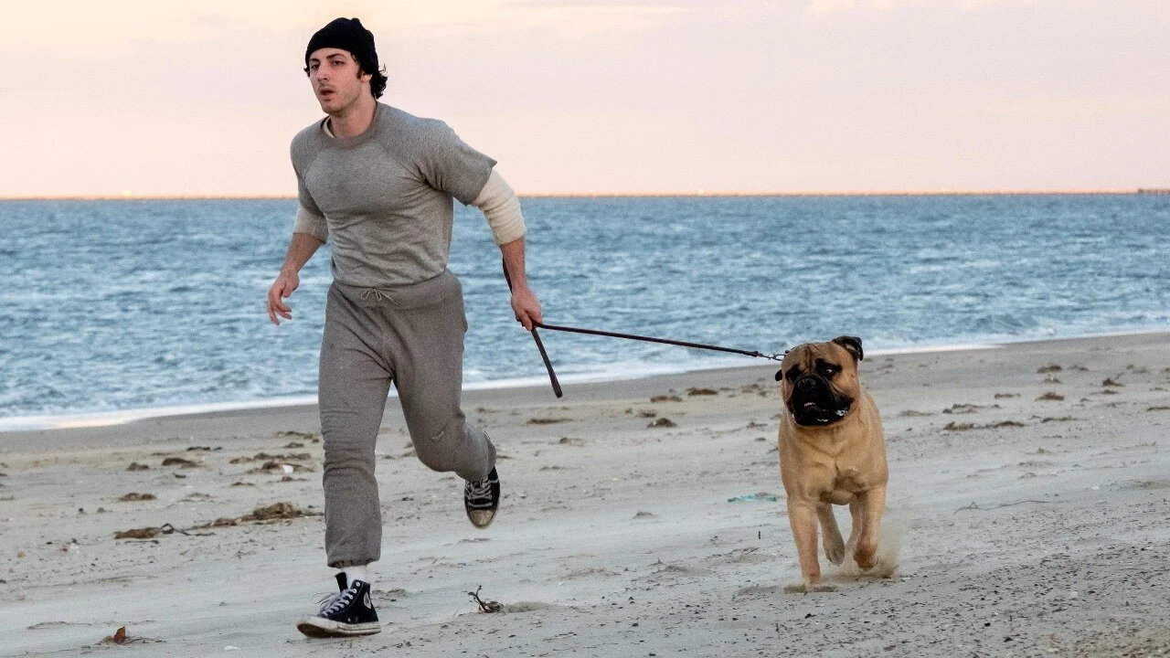 Anthony Ippolito as Sylvester Stallone runs with a dog along a beach in I Play Rocky.