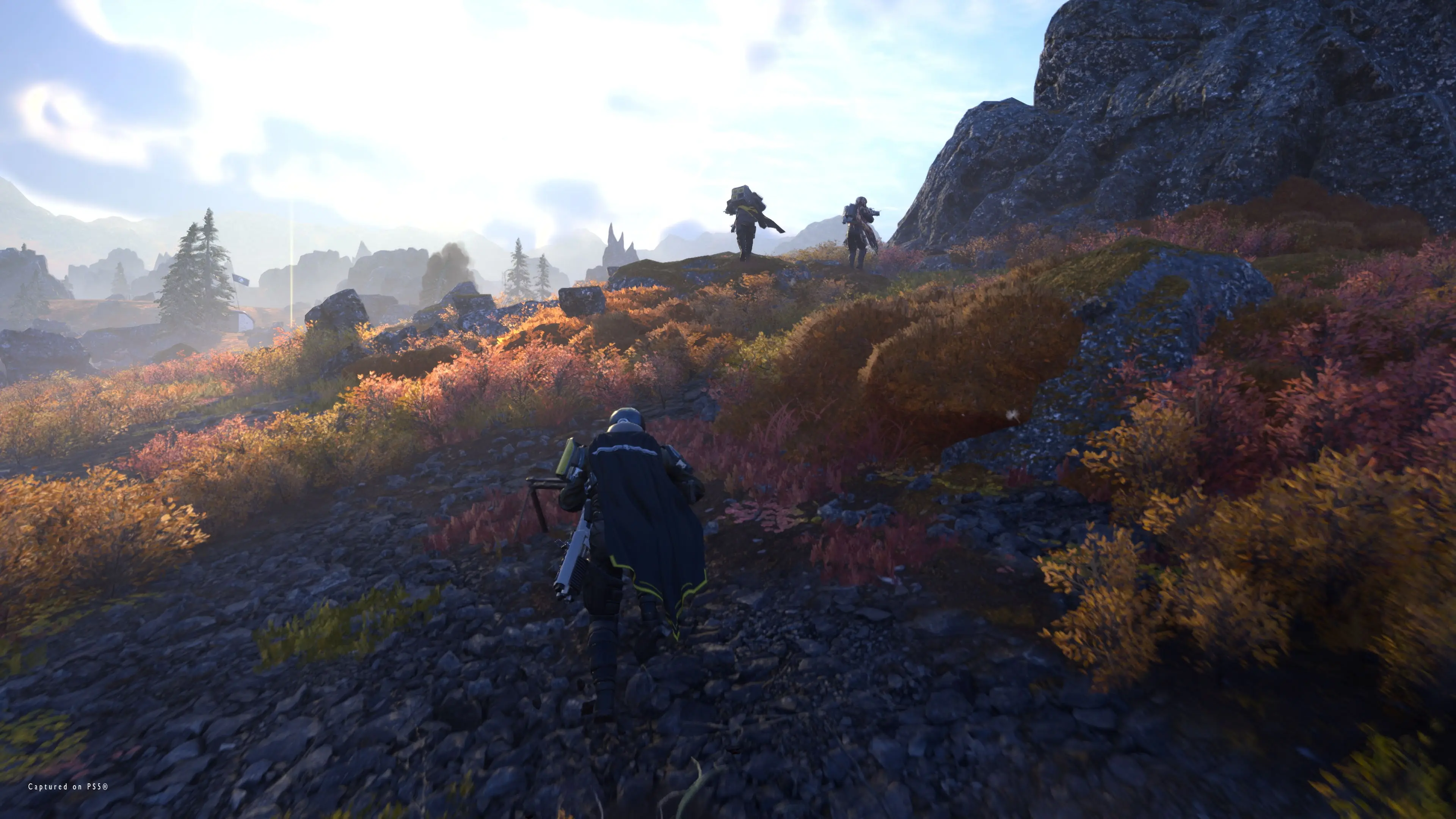 A Helldiver follows three squadmates across rocky terrain covered in orange alien plants.