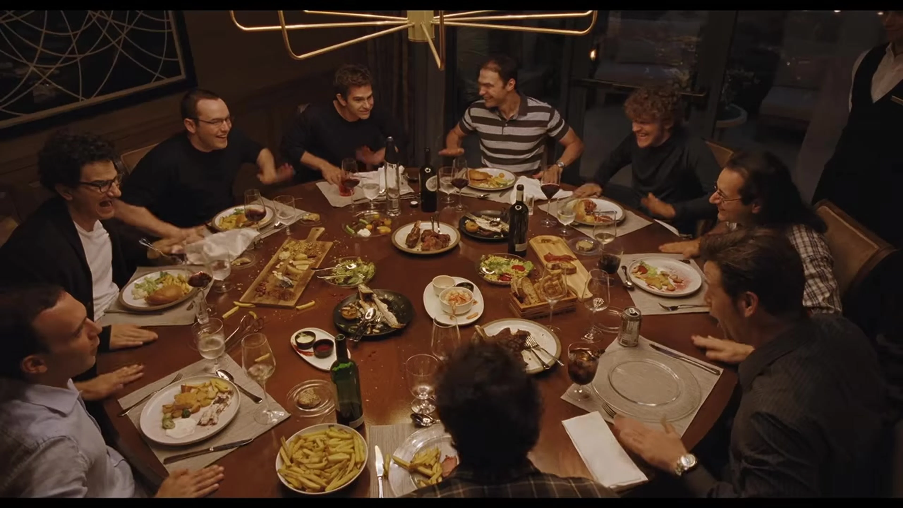 Andrew Garfield, Yura Borisov, and other cast members sit around a dinner table in Artificial.