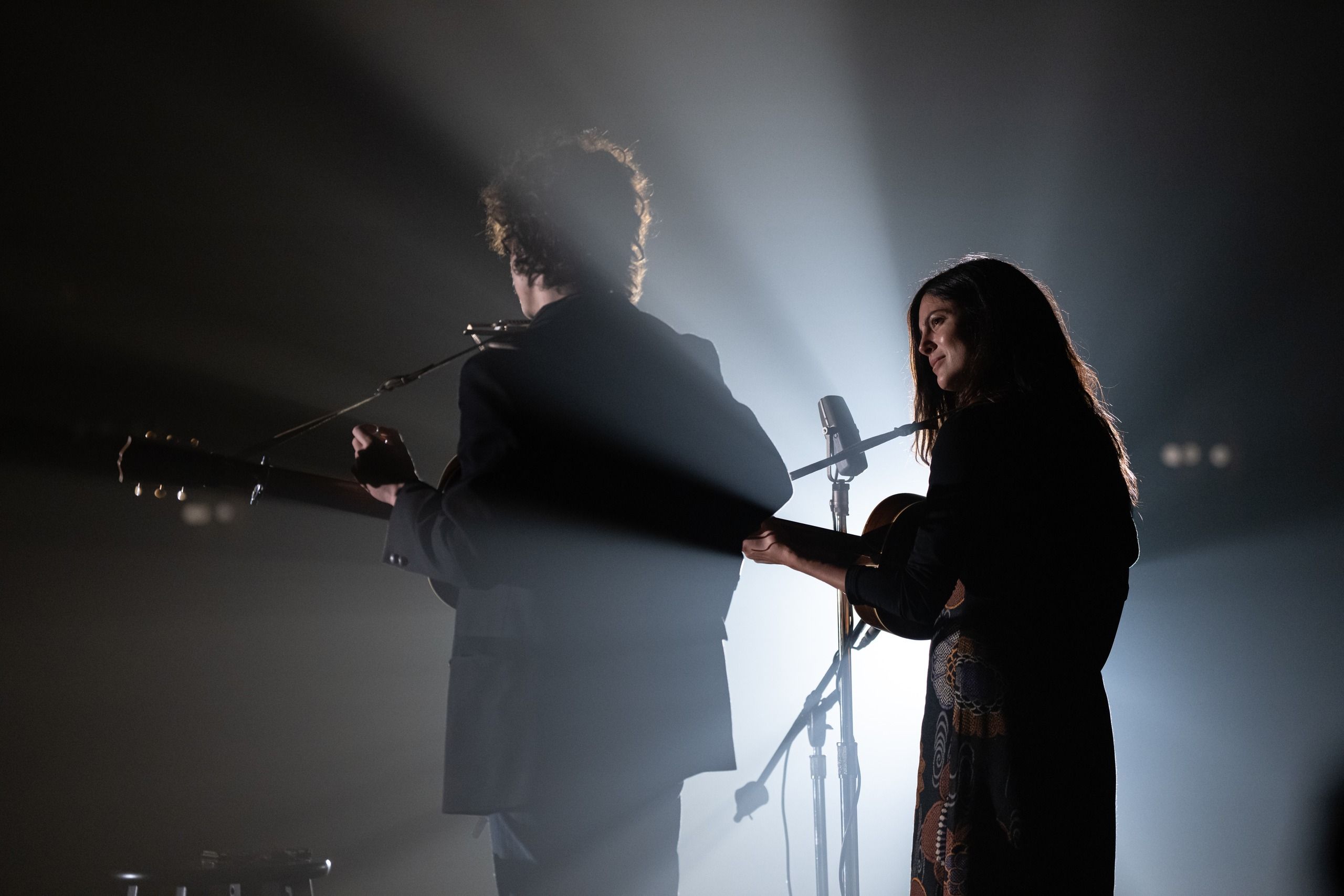 Timothée Chalamet and Monica Barbaro stand at microphones with guitars on a dimly lit stage.