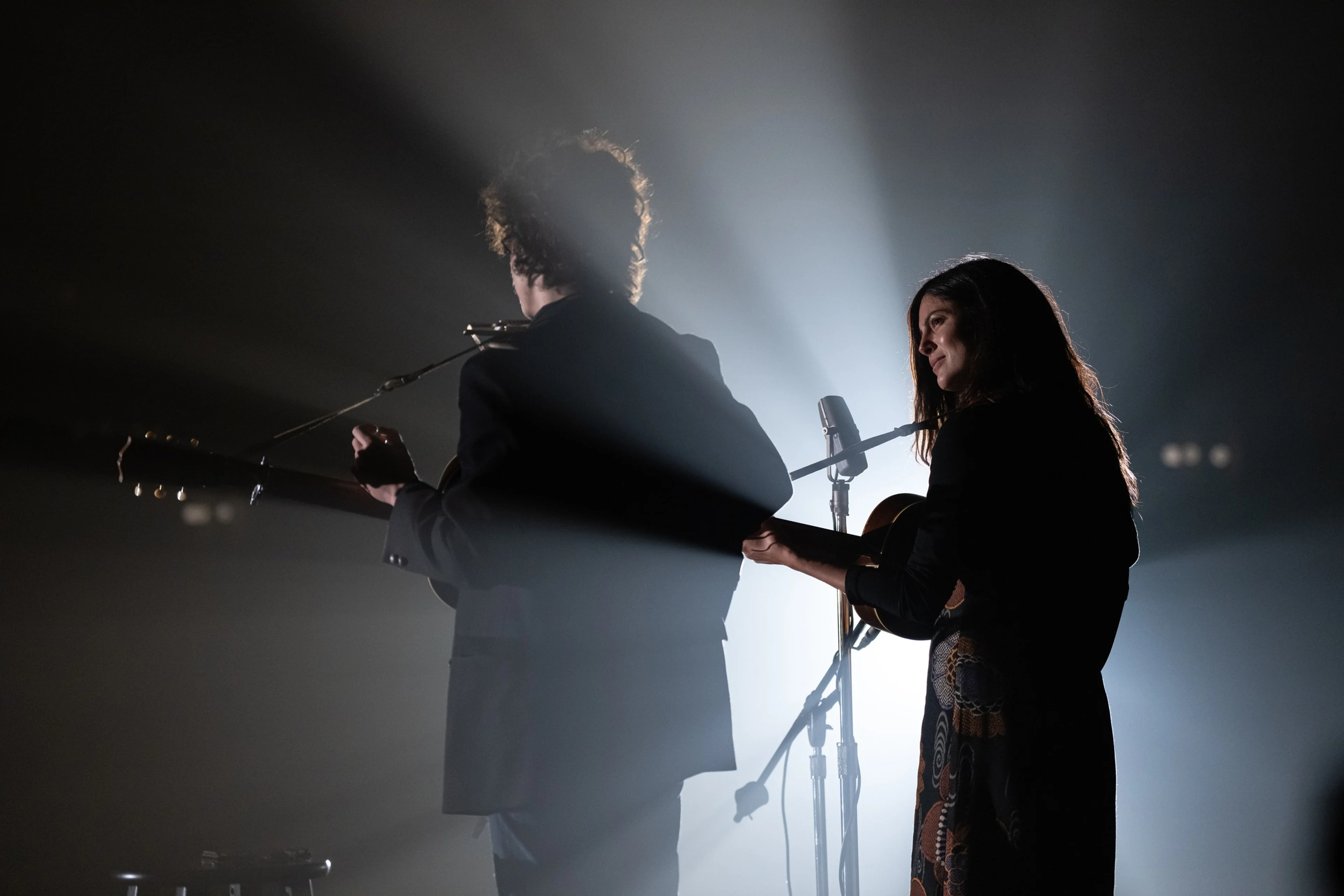 Timothée Chalamet and Monica Barbaro stand at microphones with guitars on a dimly lit stage.