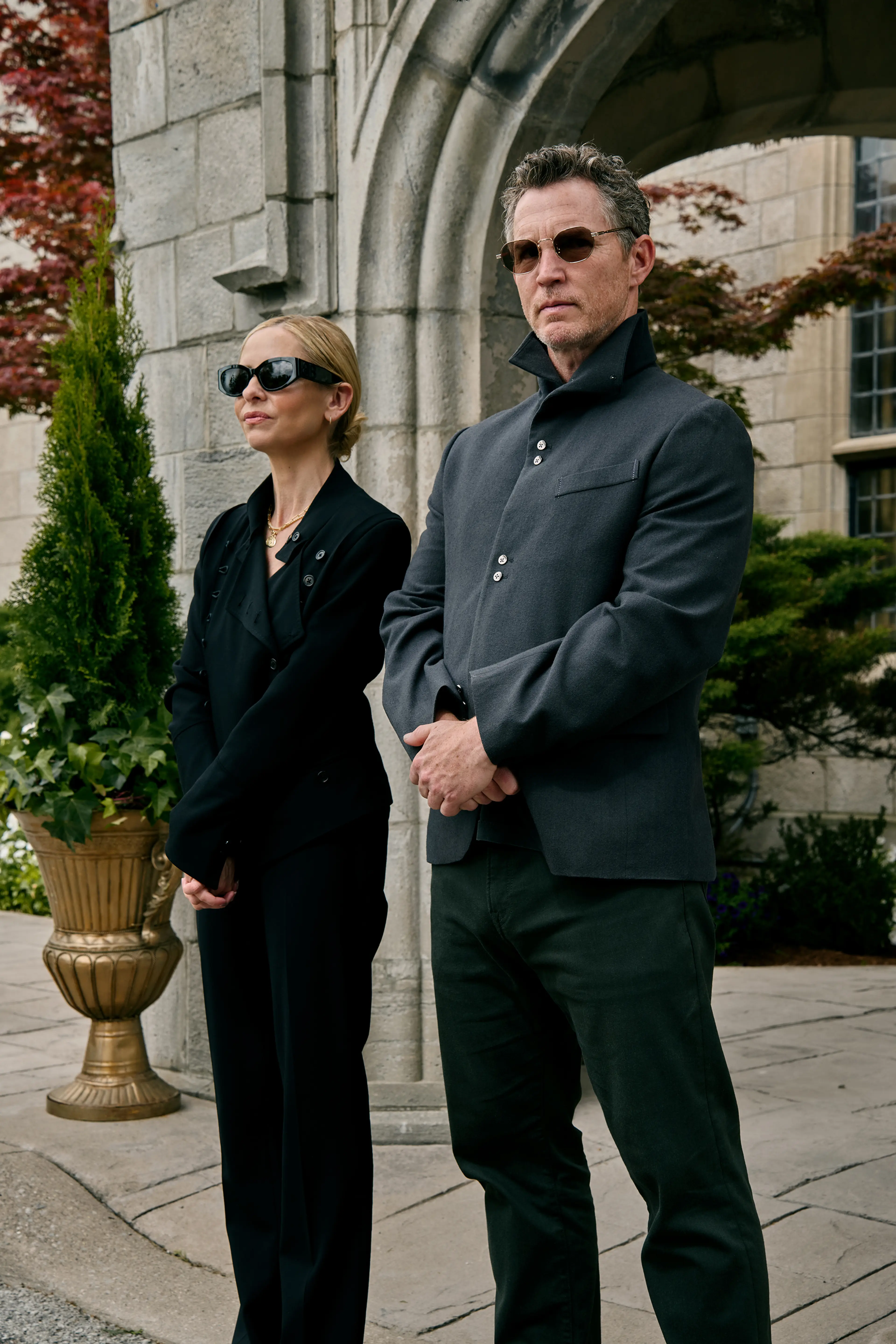 Ursula and Titus Danforth wear sunglasses and dark clothes outside a stone archway.