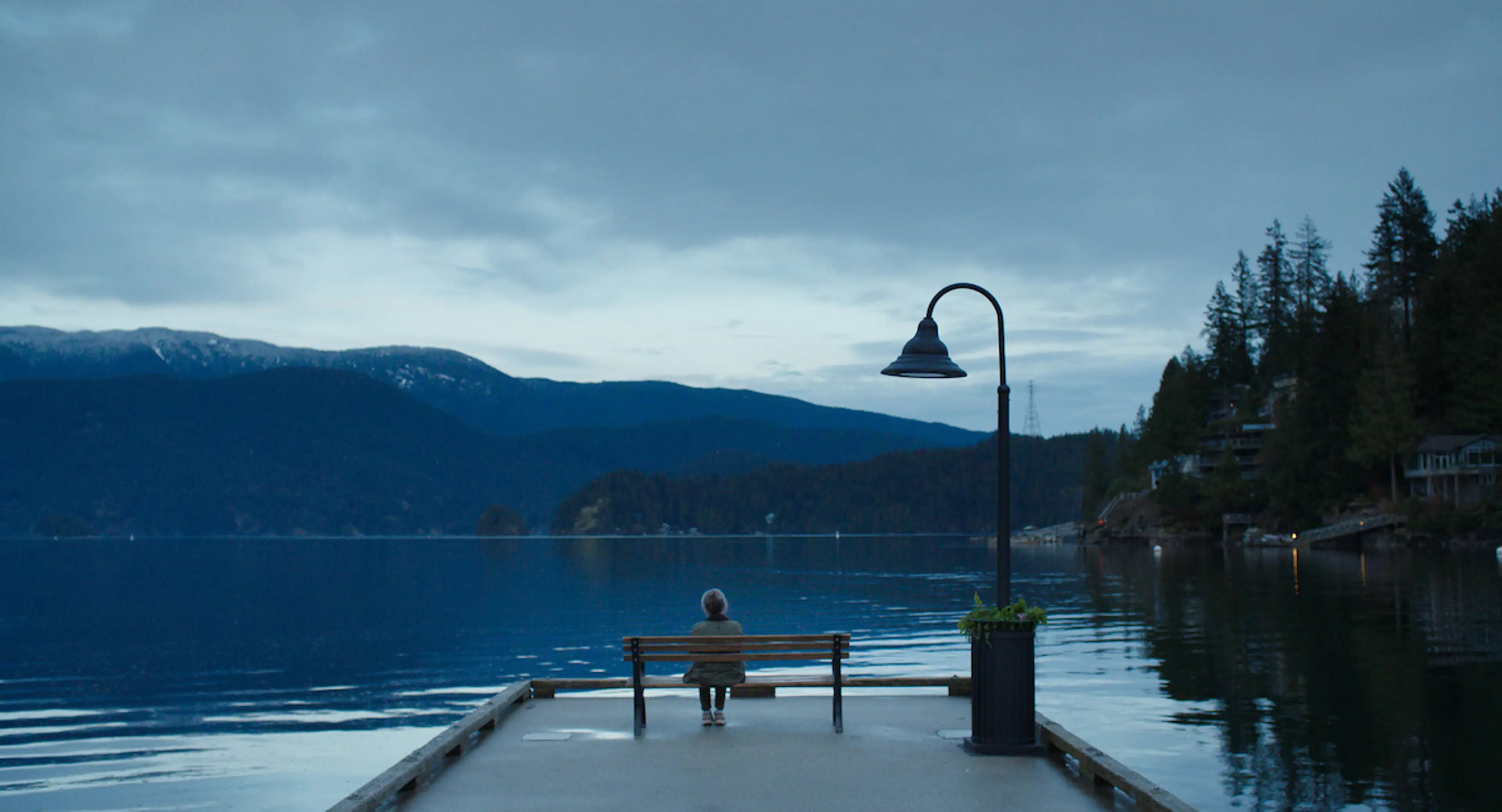 Tova sits alone on a bench at the end of a dock facing the water and mountains.