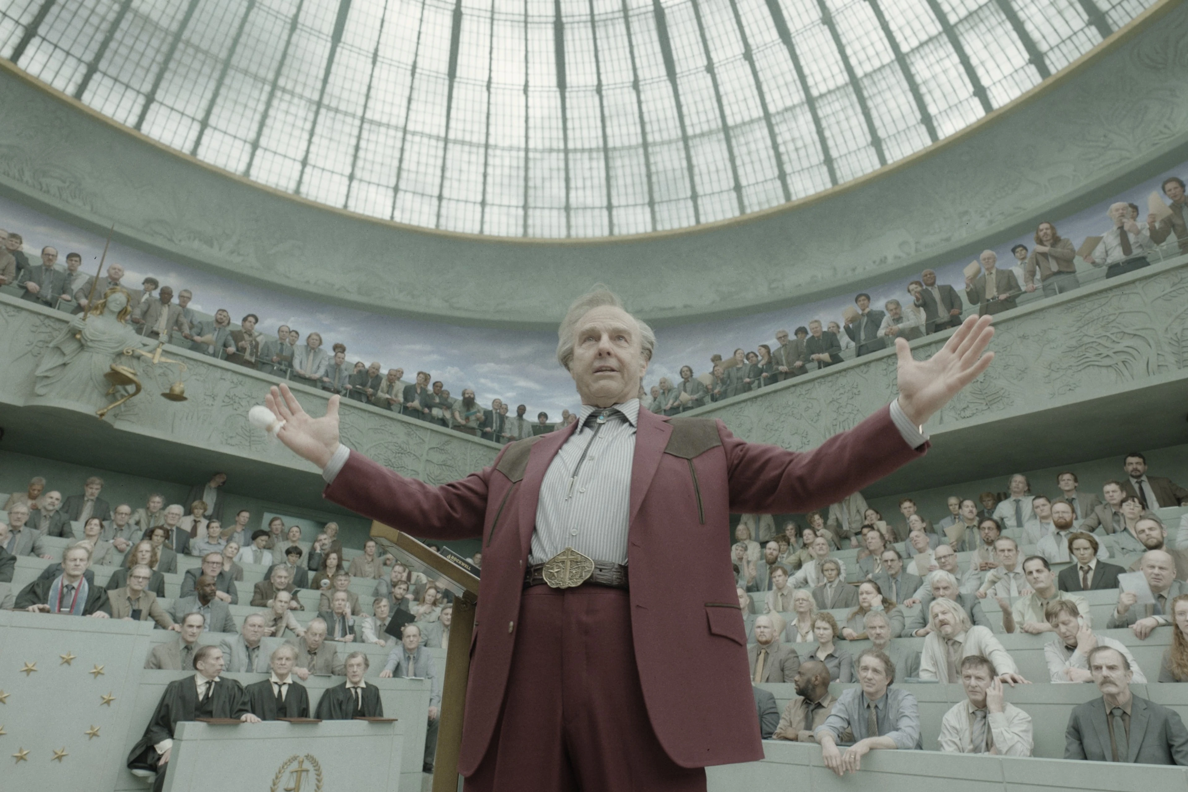 Tom Cruise in a burgundy suit addresses a crowded circular auditorium in Digger.