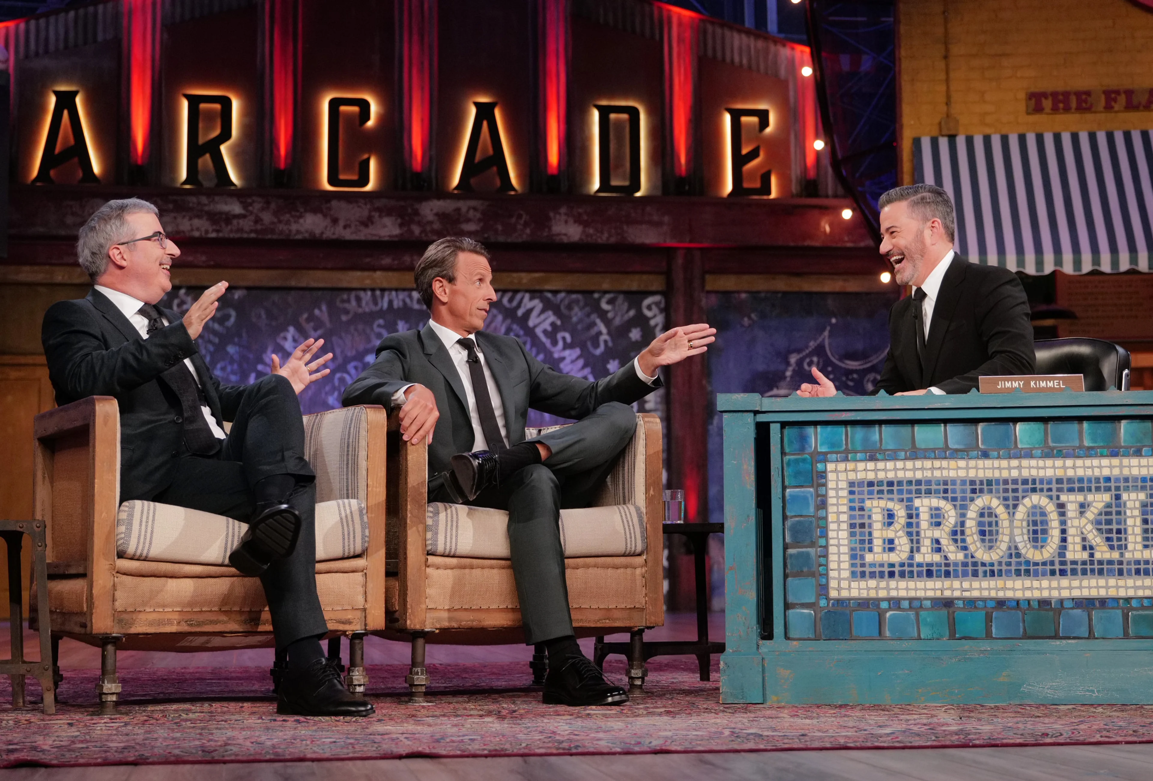 John Oliver gestures on the left while Seth Meyers and Jimmy Kimmel smile beside him.