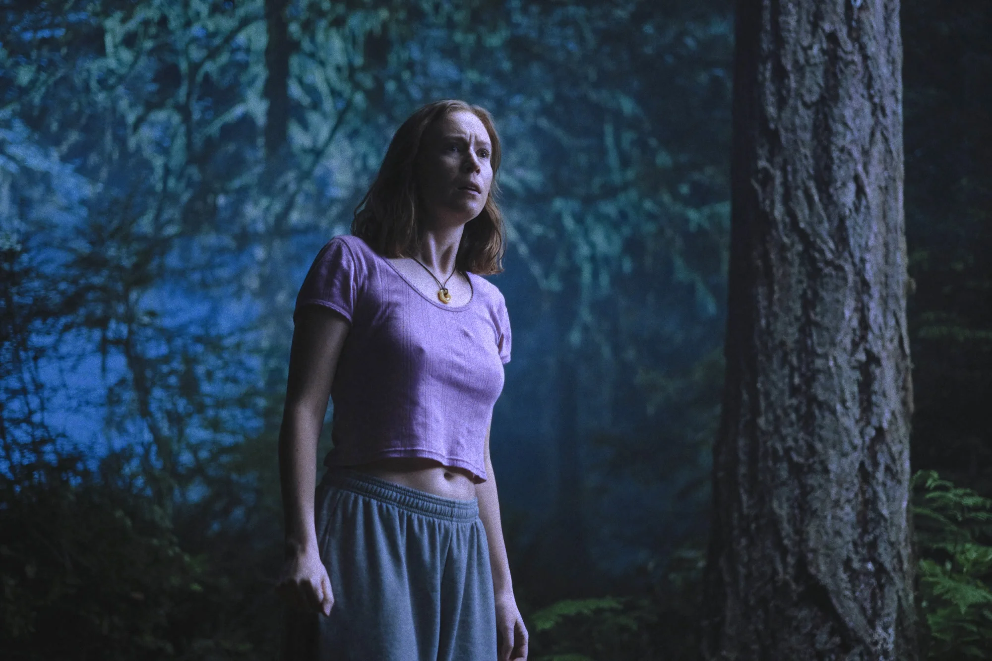 Hannah Einbinder as Kris stands beside a large tree in a blue-lit forest at night.