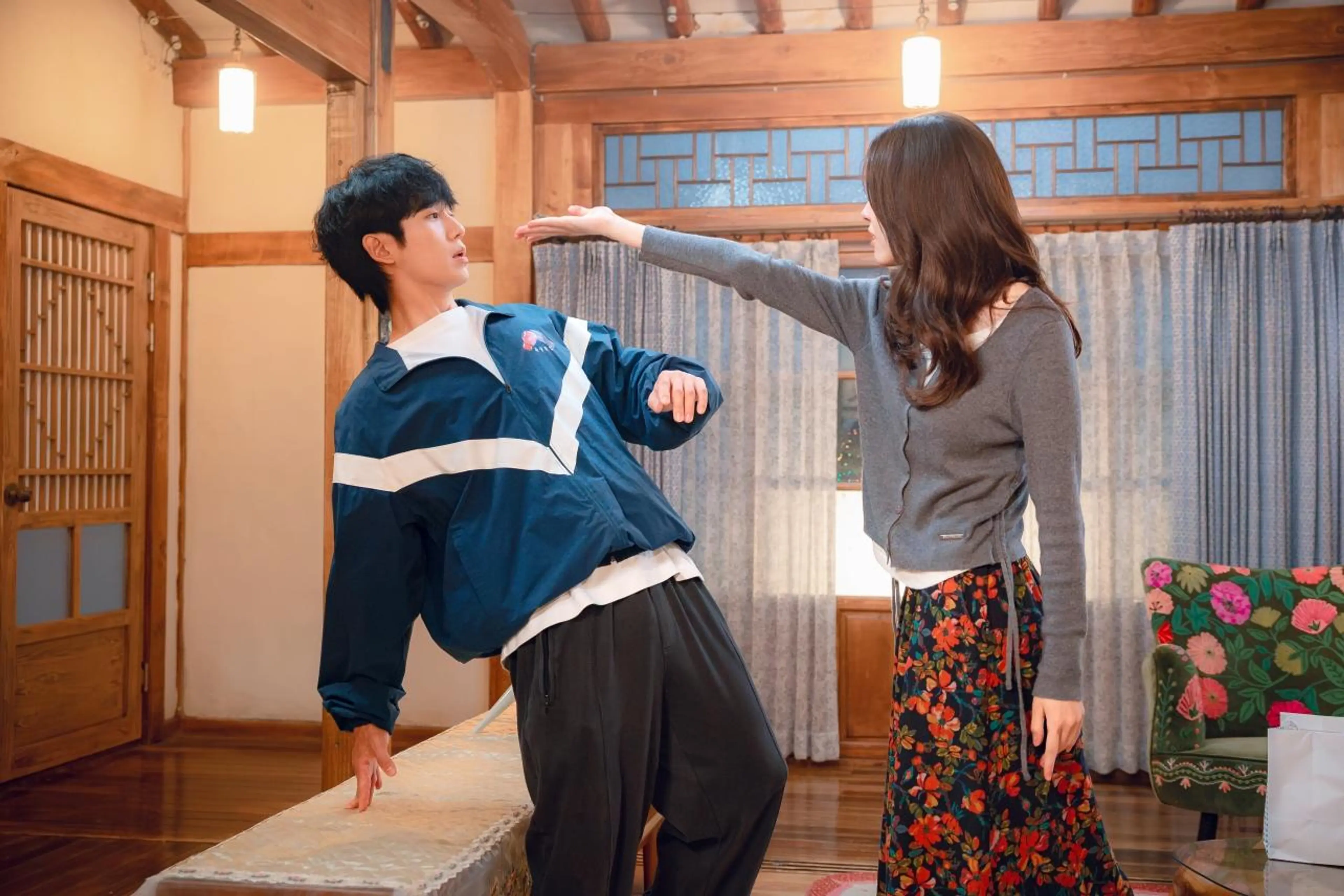 Ha Young reaches towards Jung Hae-in’s face as he leans backwards beside a bed in a traditional wooden house.