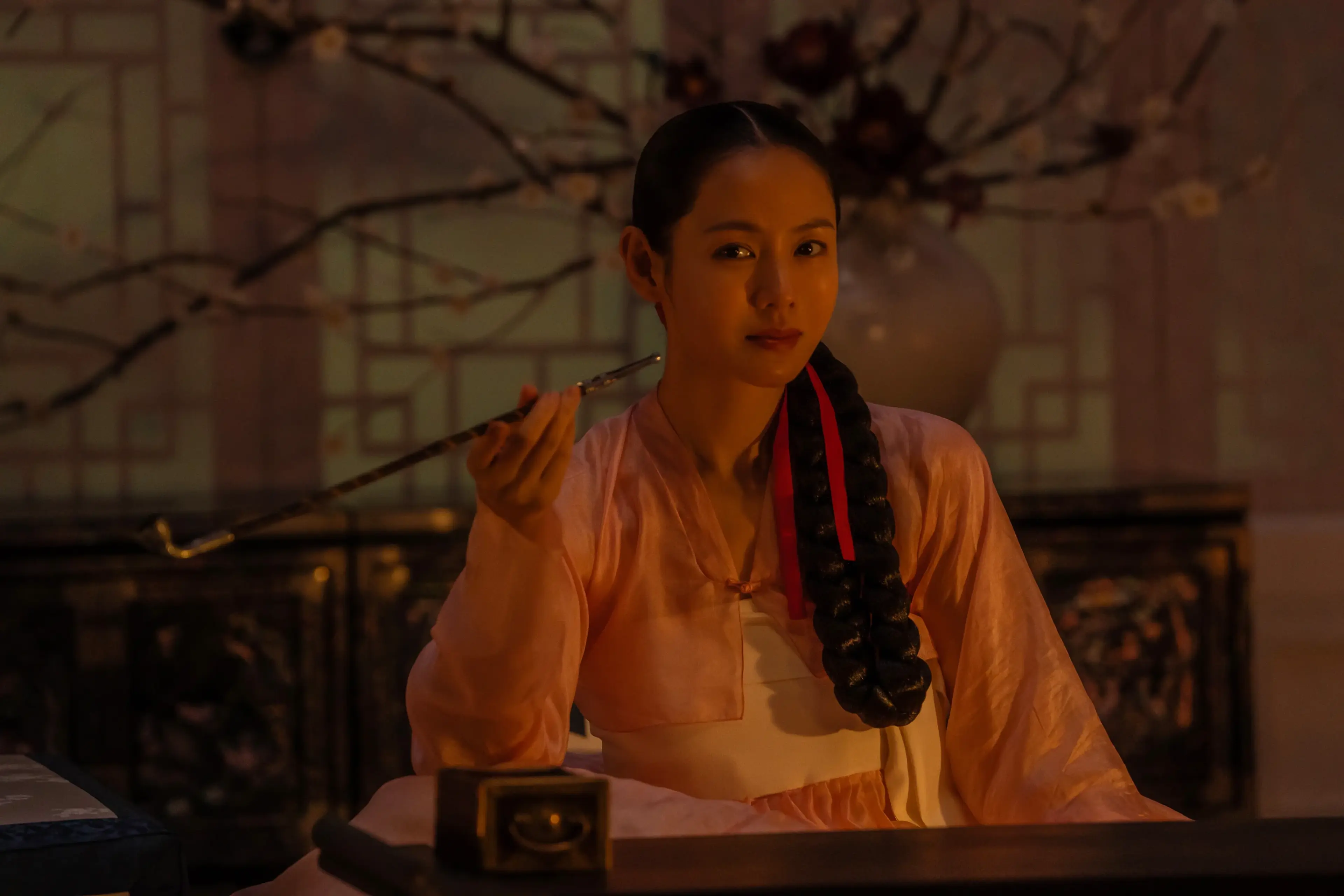 Lady Cho holds a long pipe while seated beside a table in a dim room.