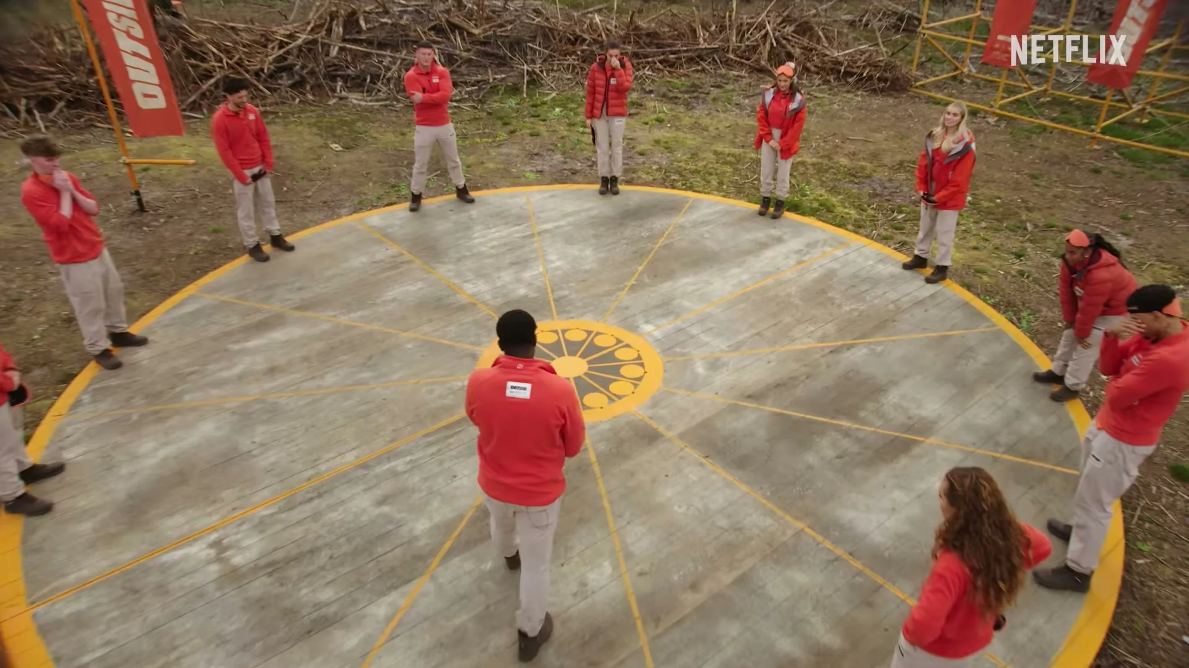 Outside contestants gather around a circular challenge area in the forest.