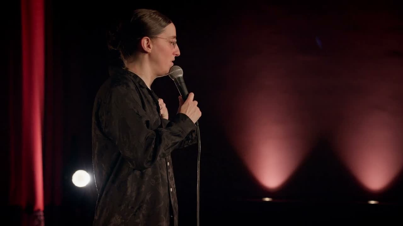 Robby Hoffman faces the audience with one hand raised near her microphone in Wake Up.