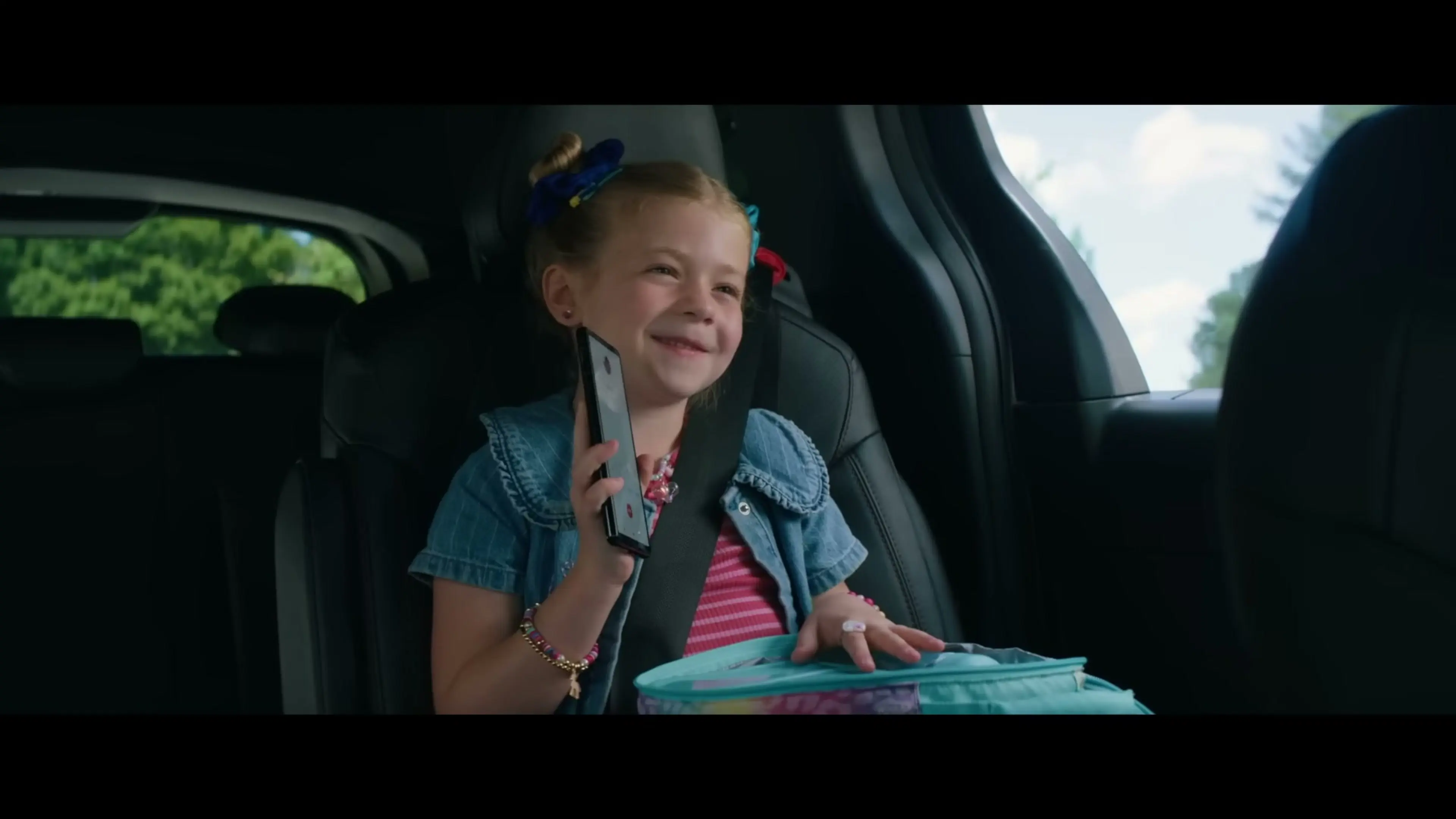 A young girl sits in the back of a car holding a phone.
