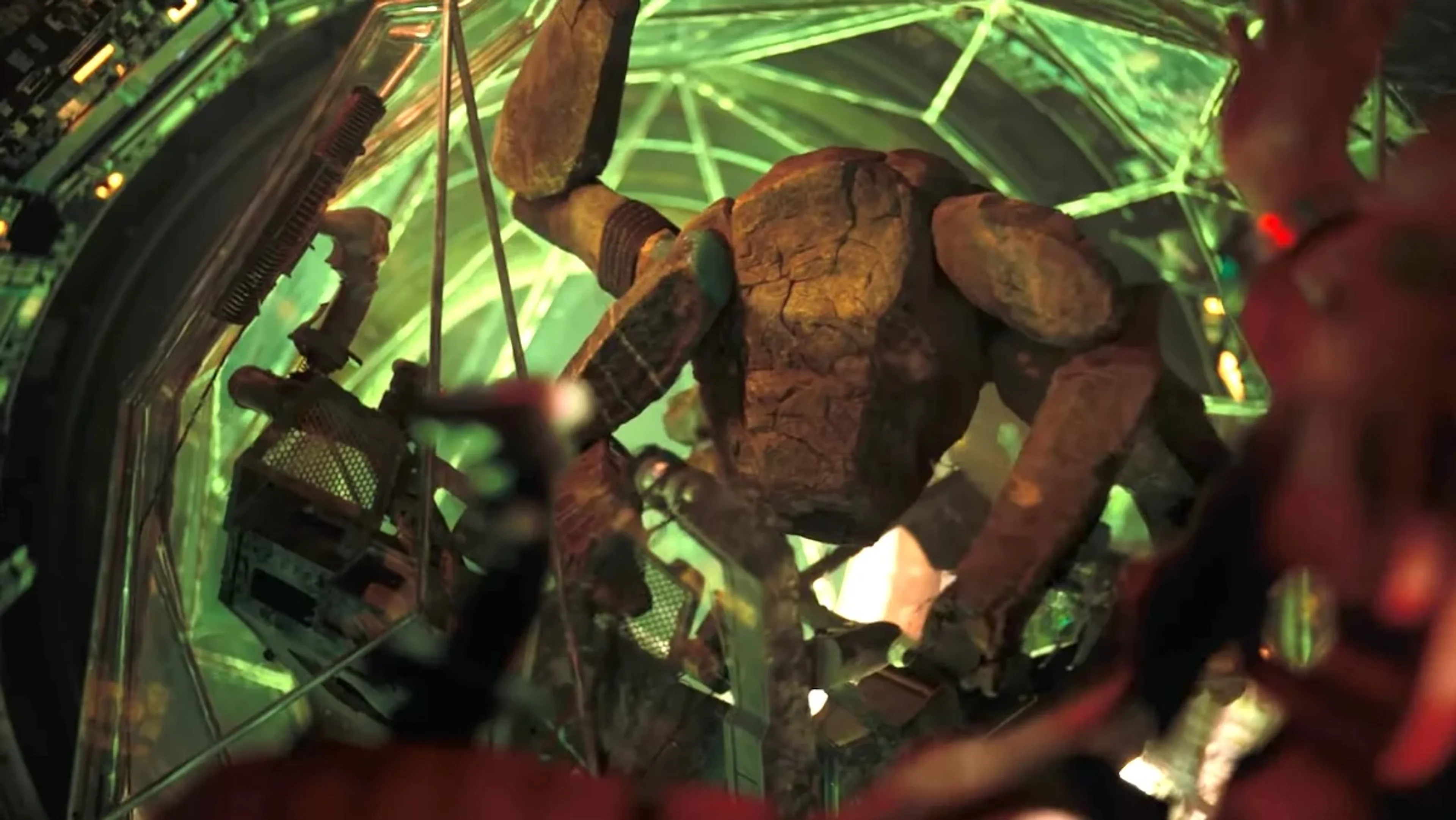 Rocky, a multi-limbed alien with a rough brown body, raises an arm inside a green-lit chamber.