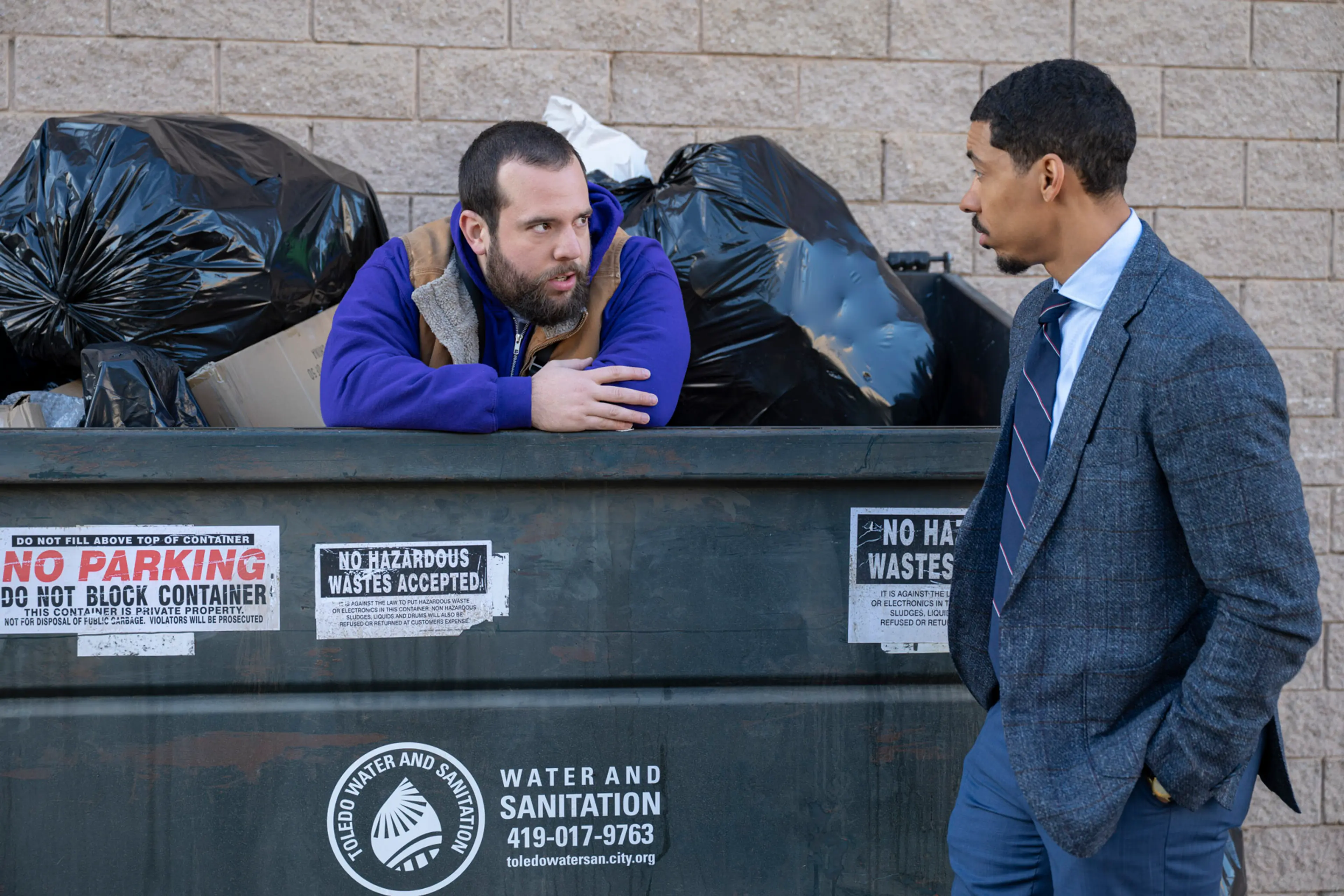 Travis leans out of a dumpster while Detrick stands next to it.
