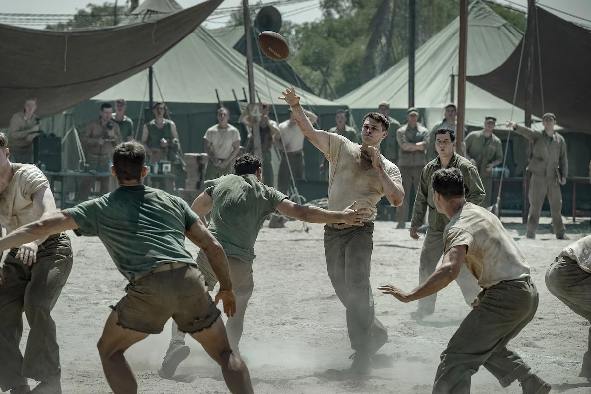 Nicholas Galitzine as John McLaughry carries a football while Marines watch from the sideline in The Mosquito Bowl.