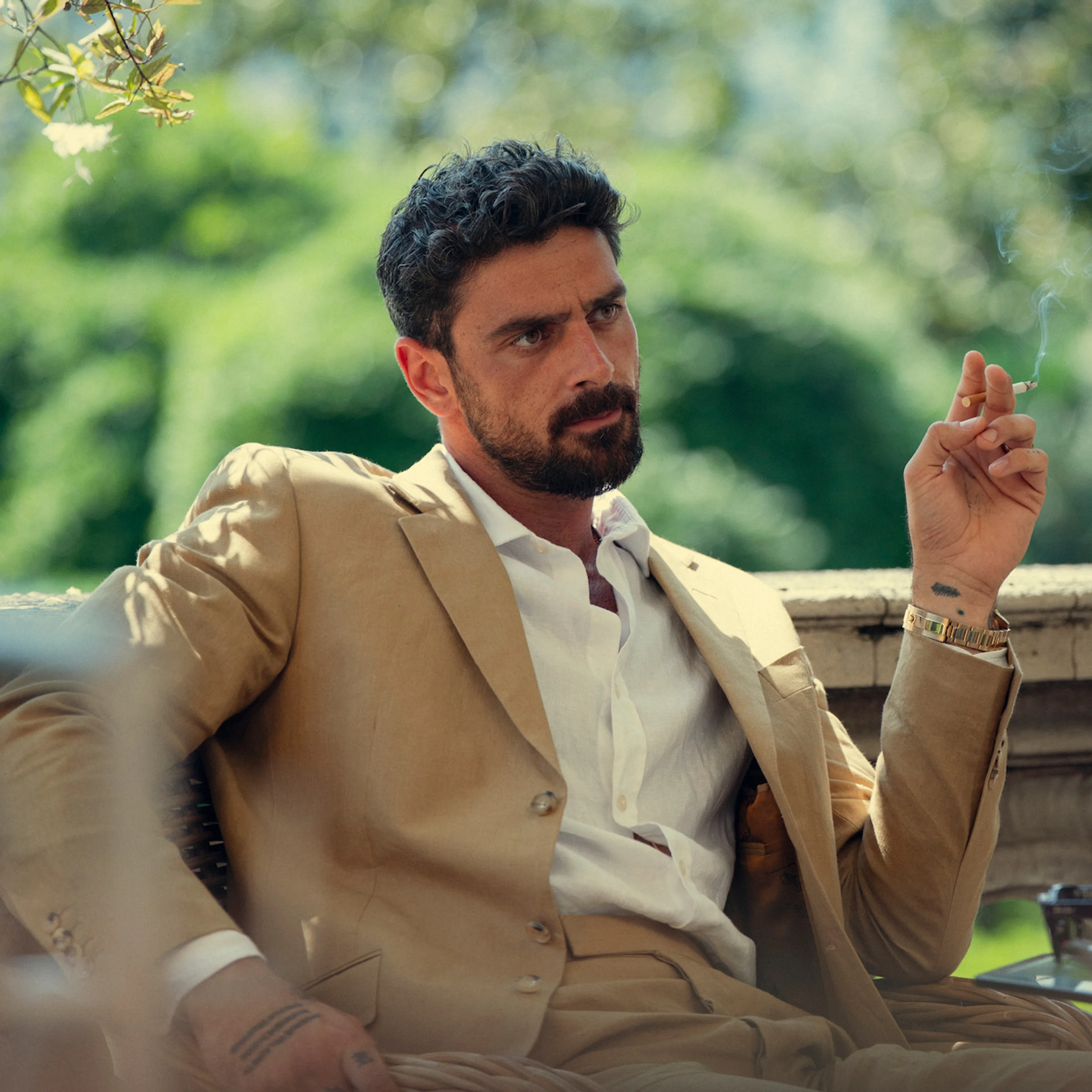 Michele Morrone as Cico Maldini, seated outdoors in a tan suit in The Gentlemen Season 2.