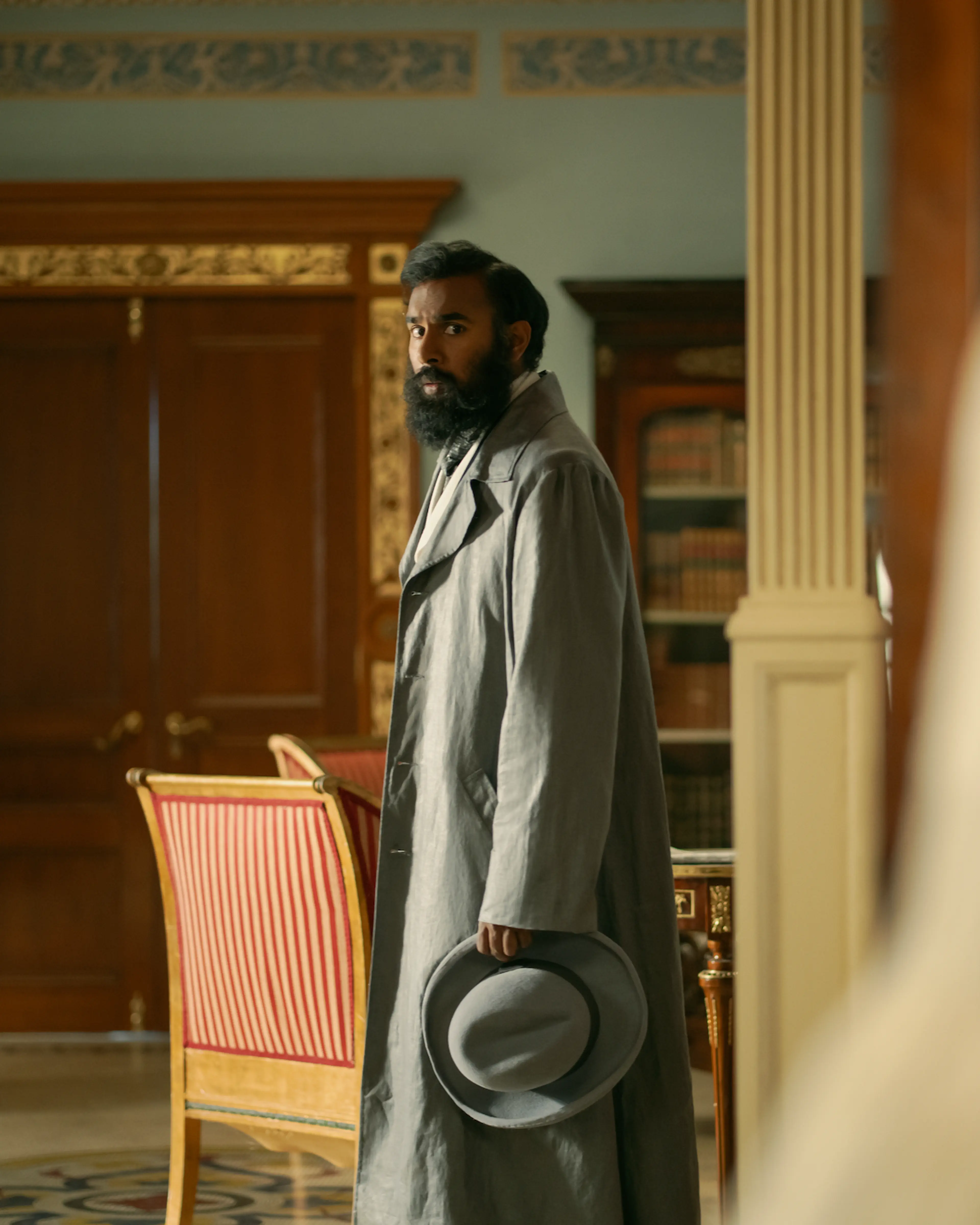 Himesh Patel as Dr. Watson stands in a furnished room wearing a long gray coat and holding a hat.