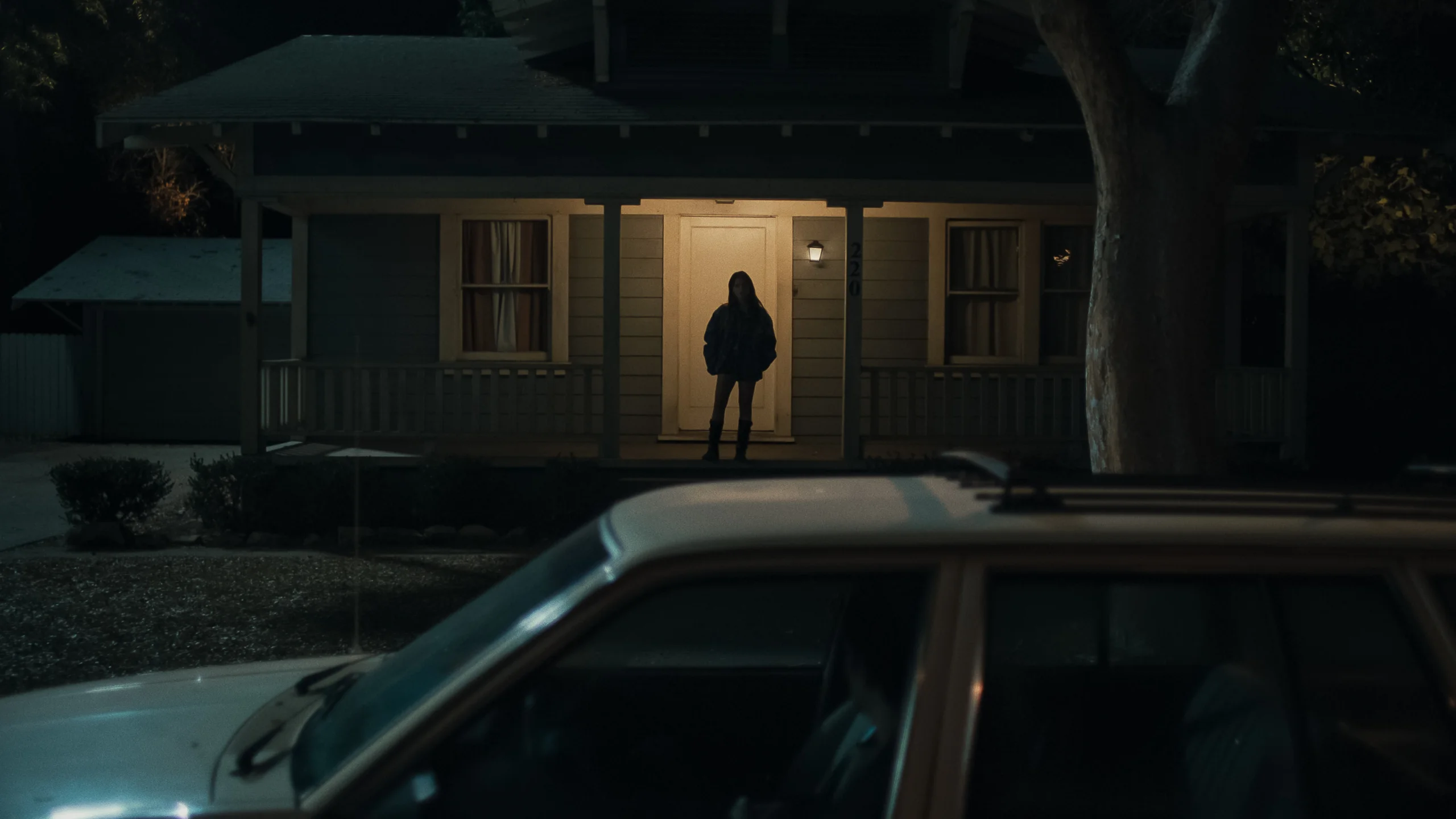 Nikki stands on a lit porch at night, with a car in the foreground.