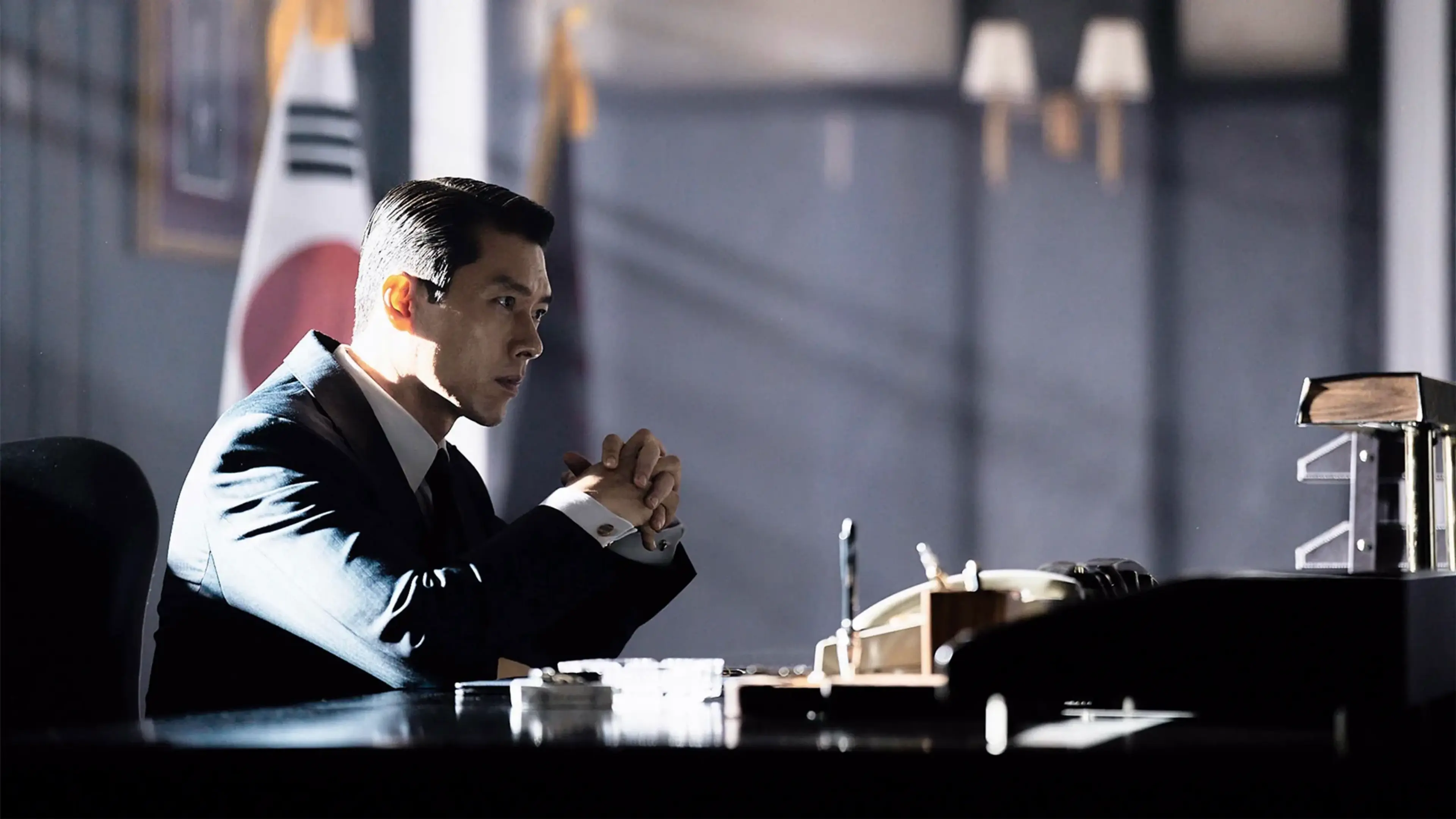 Hyun Bin as Ki-tae sits at a desk with his hands clasped beside a Korean flag.