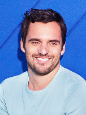 Portrait of Jake Johnson smiling in a pale blue top against a blue background.