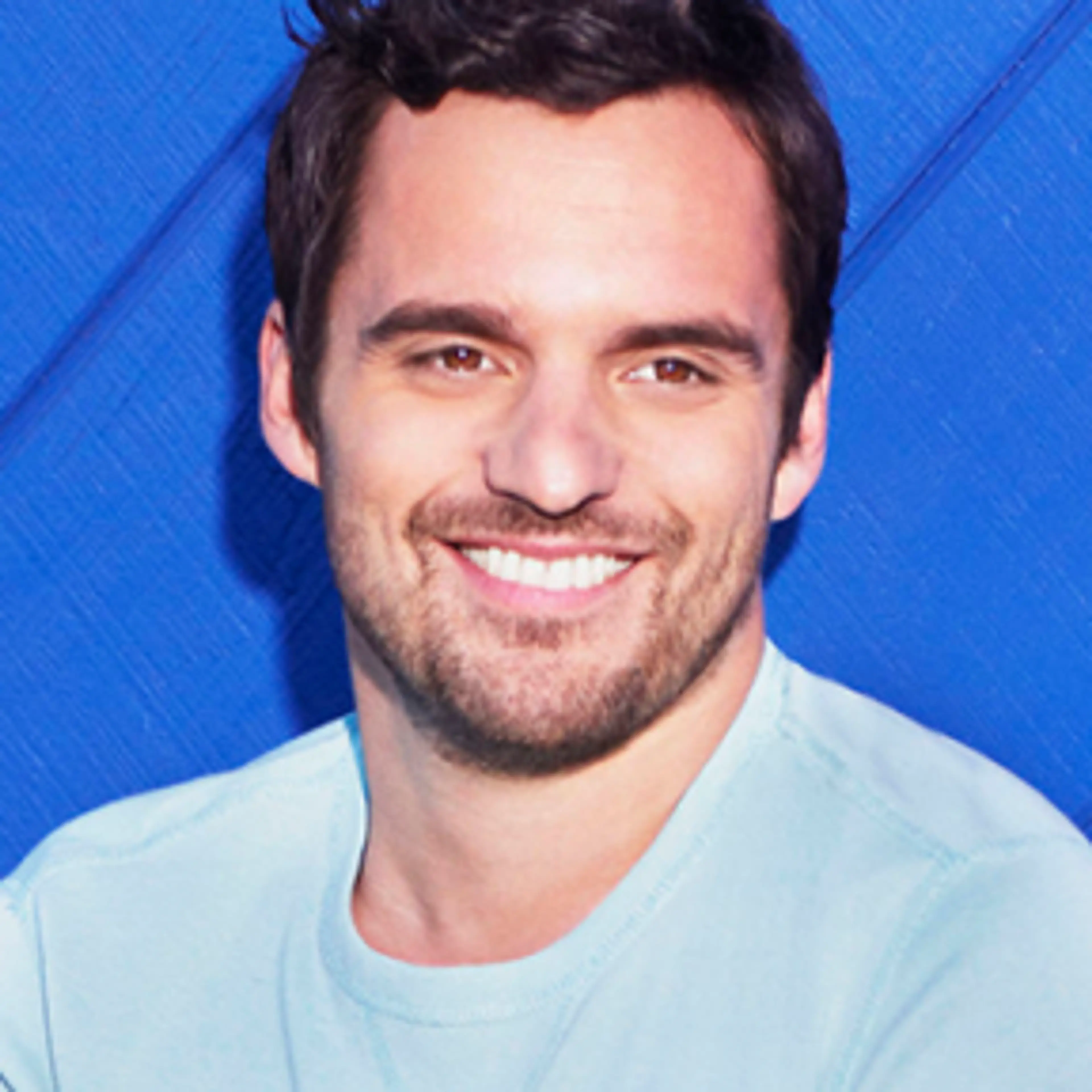 Portrait of Jake Johnson smiling in a pale blue top against a blue background.