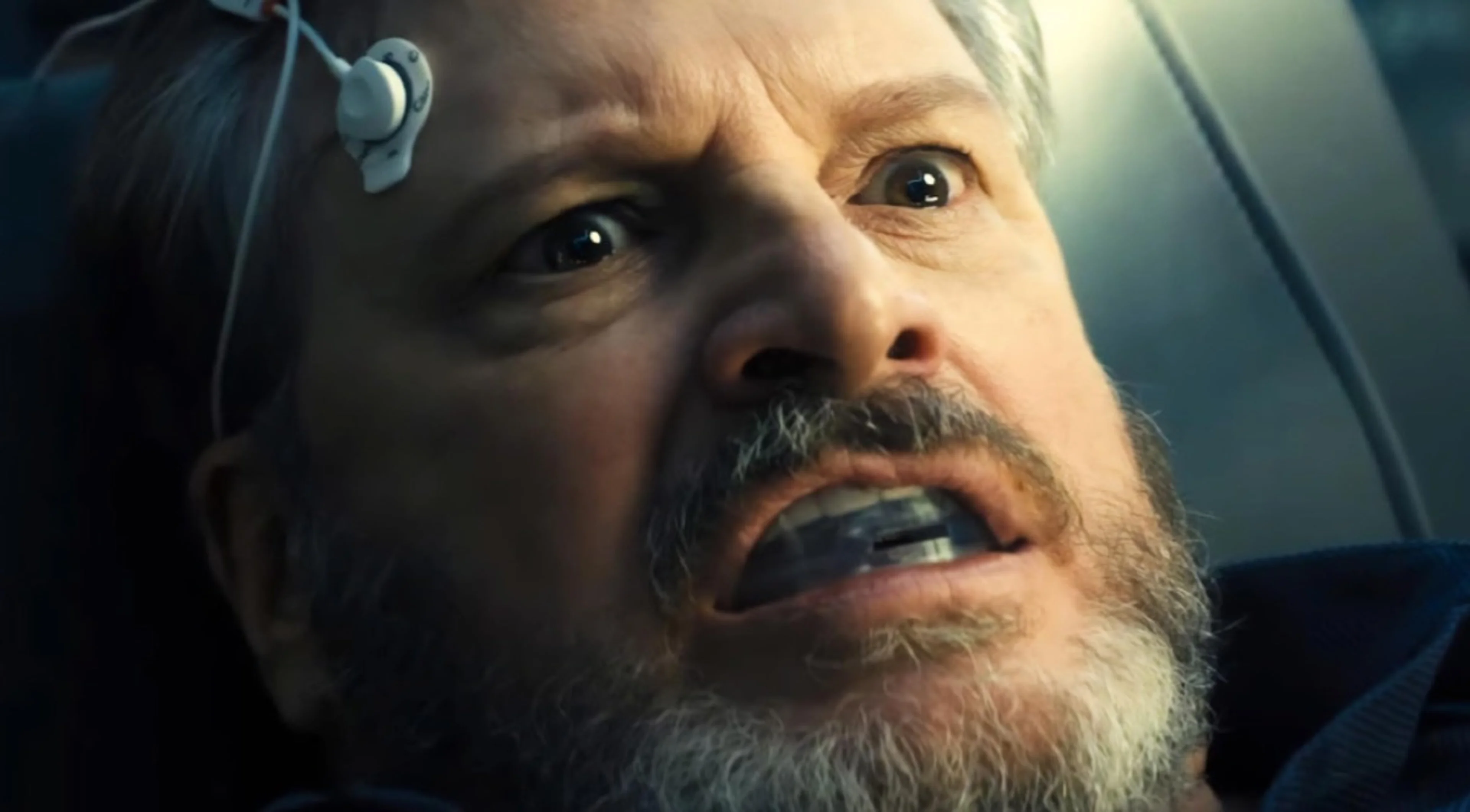 Colin Firth as Noah Scanlon wears a device beside his temple.