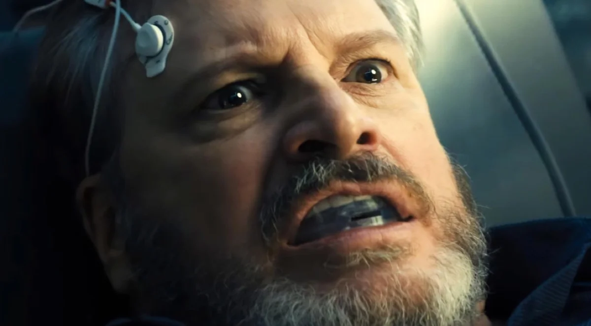 Colin Firth as Noah Scanlon wears a device beside his temple.